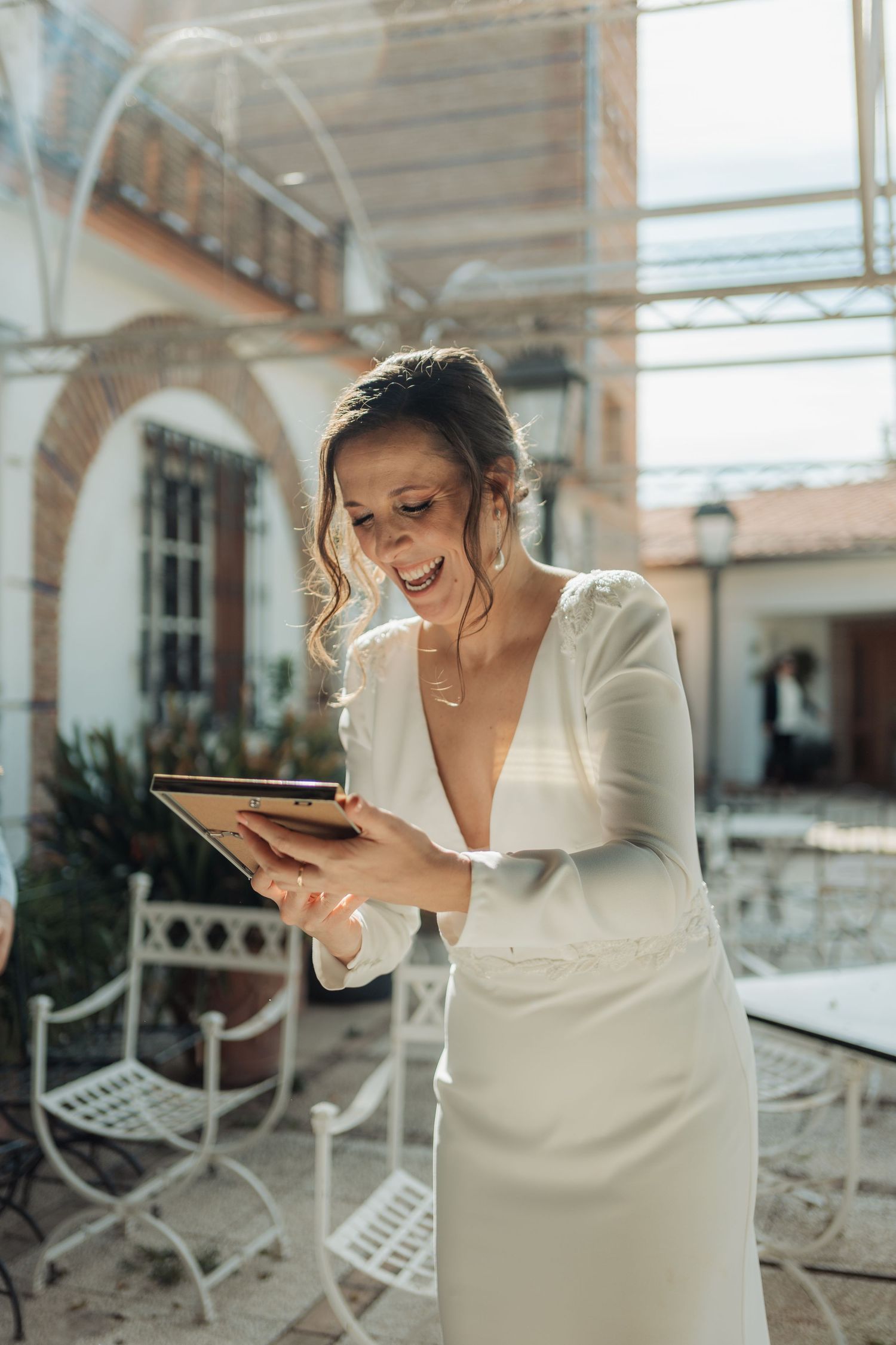 Laughing bride in white V-neck gown holding a tablet in a bright courtyard setting.
