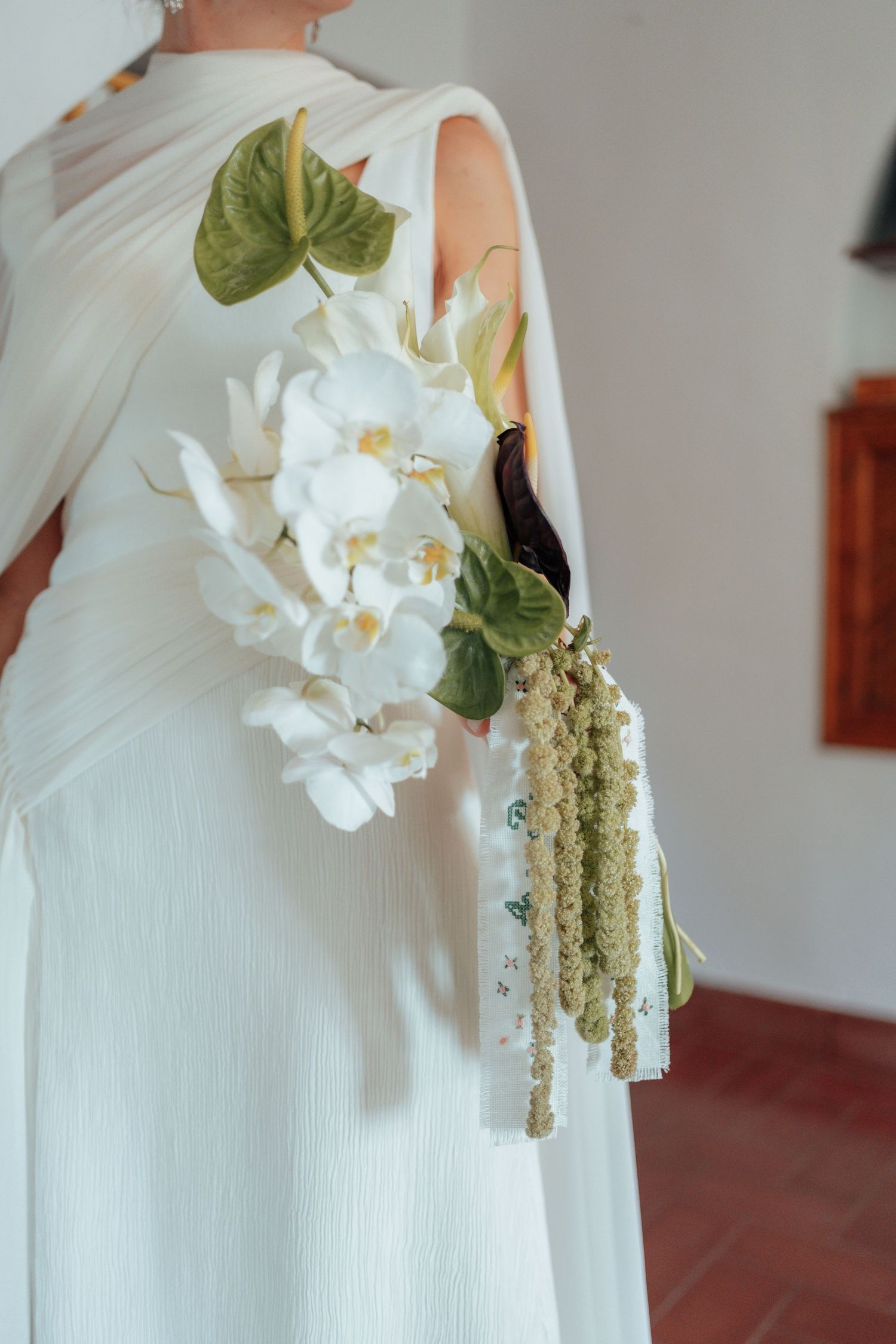 Bride in white dress holding elegant bouquet of white orchids and tropical green leaves from behind.