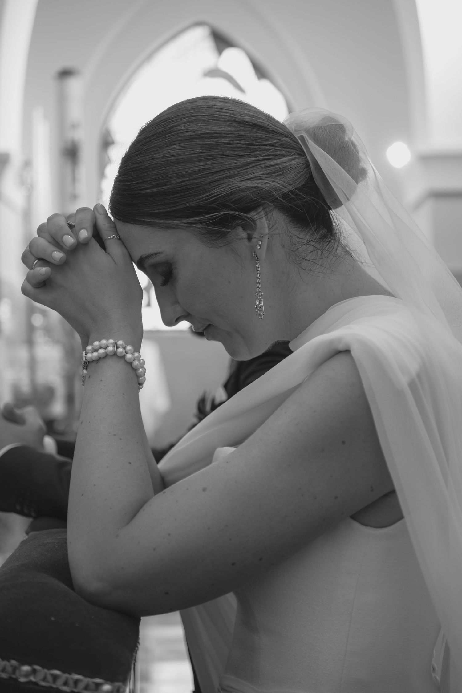Black and white photo of bride in veil praying with clasped hands during church wedding ceremony.
