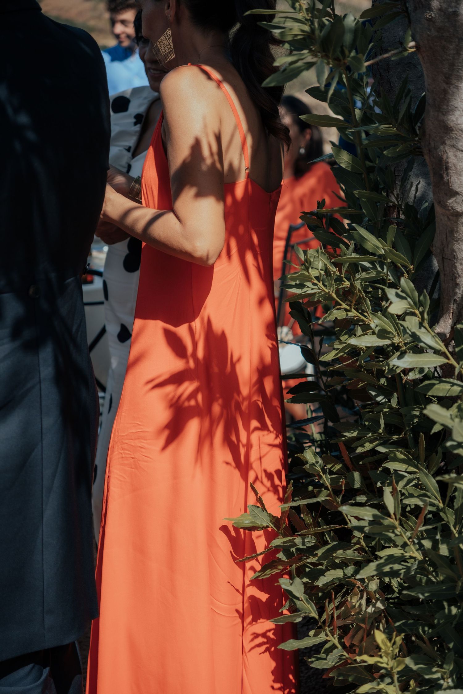 Woman in elegant orange sleeveless maxi dress standing near green foliage in dappled sunlight.
