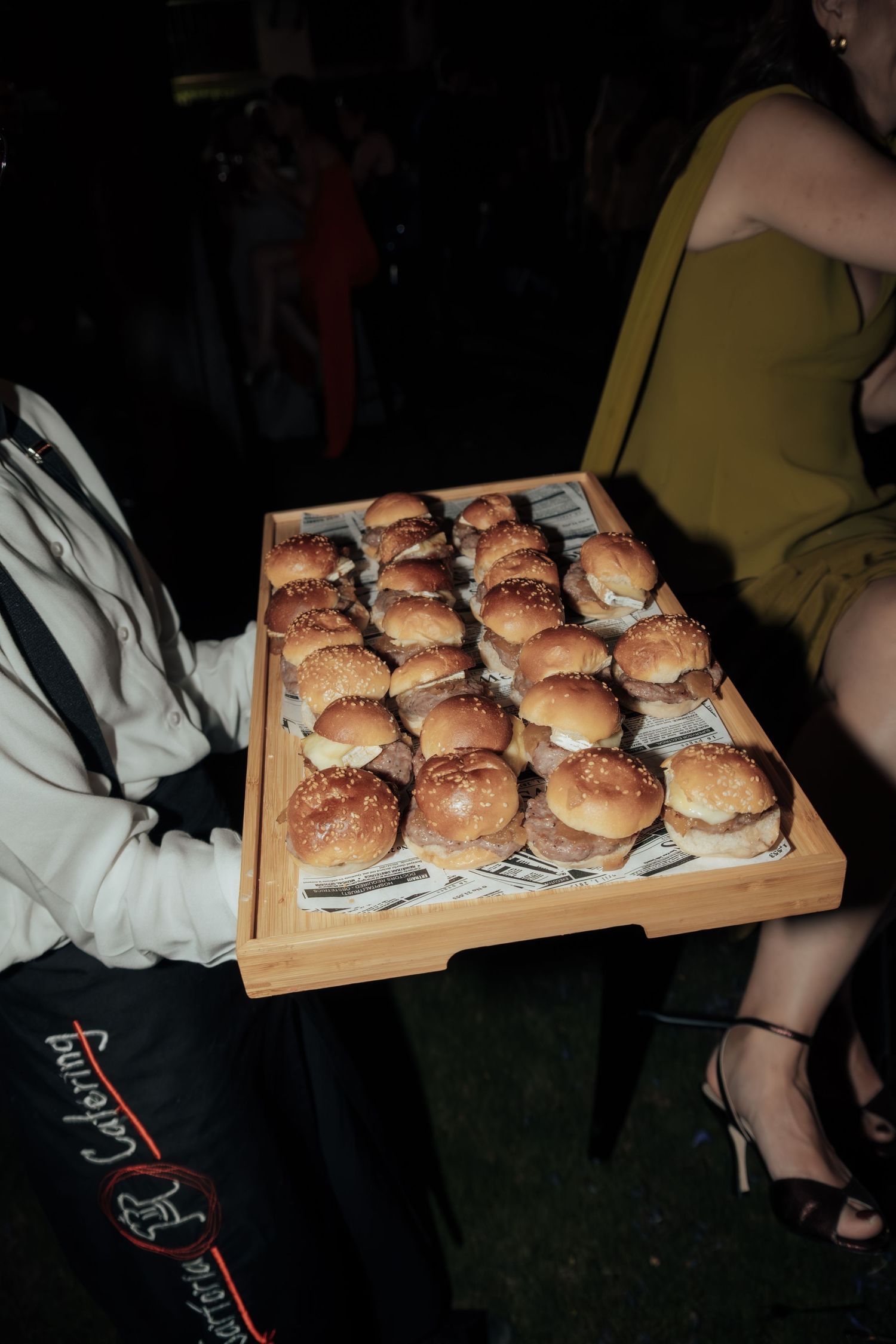 Server presenting a wooden tray filled with mini slider burgers at an outdoor evening party.