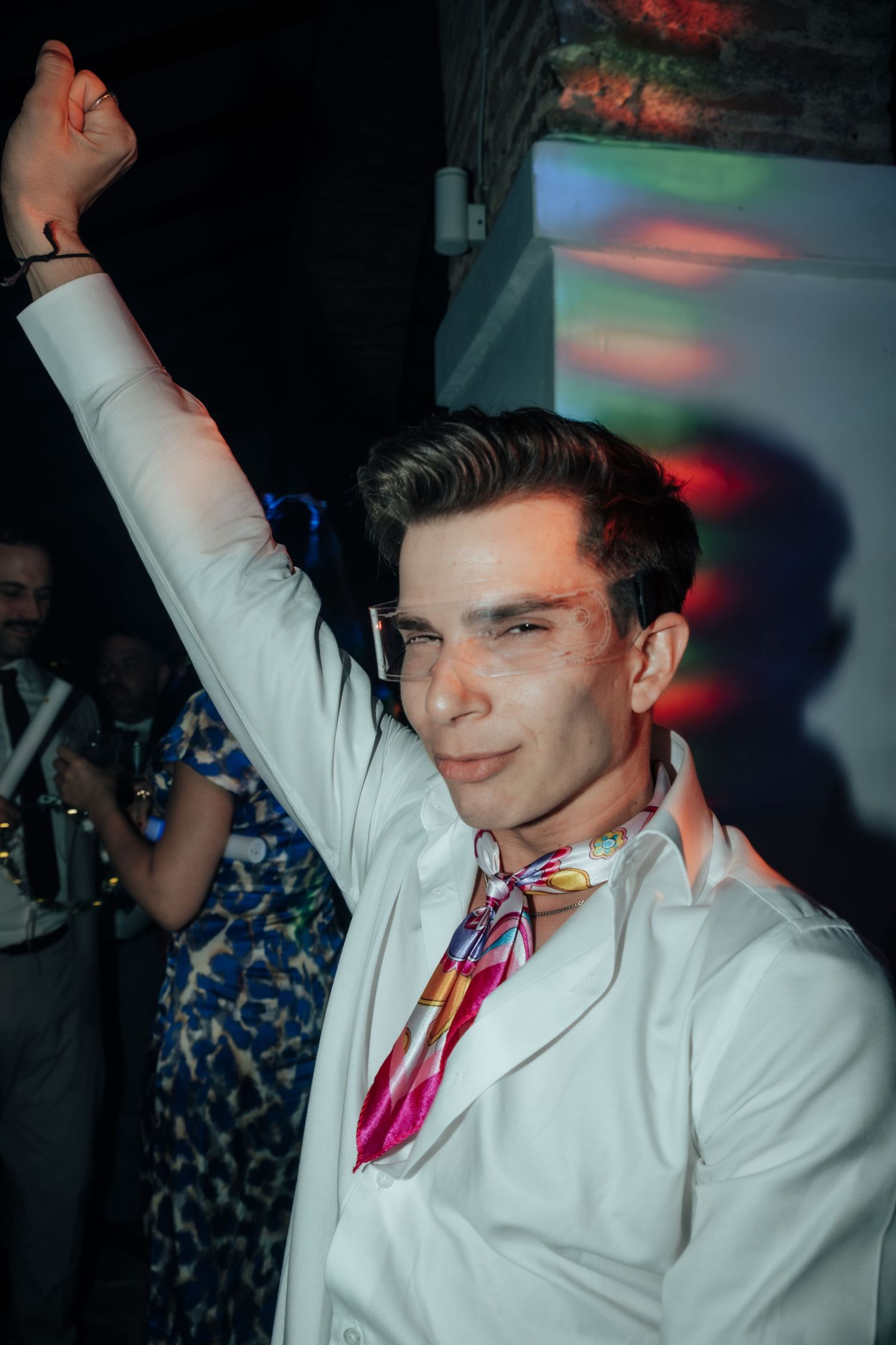 Stylish man in white jacket with colorful scarf raising arm while dancing at vibrant party.