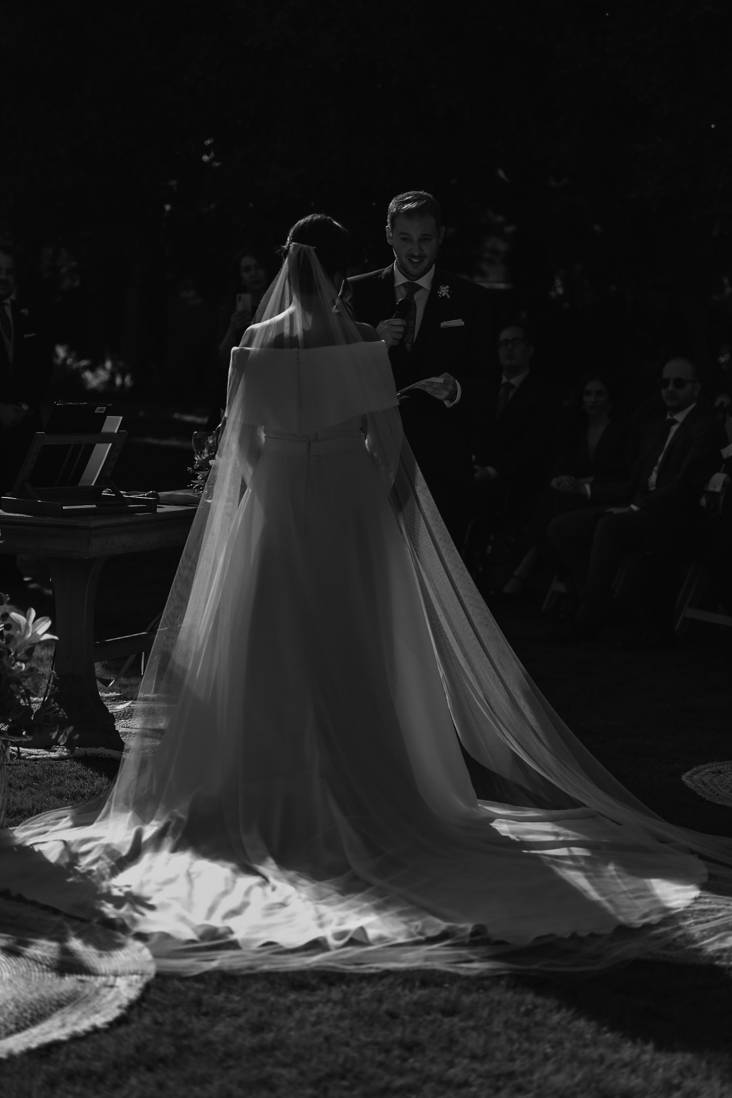 Black and white photo of bride in flowing veil and gown during outdoor wedding ceremony.
