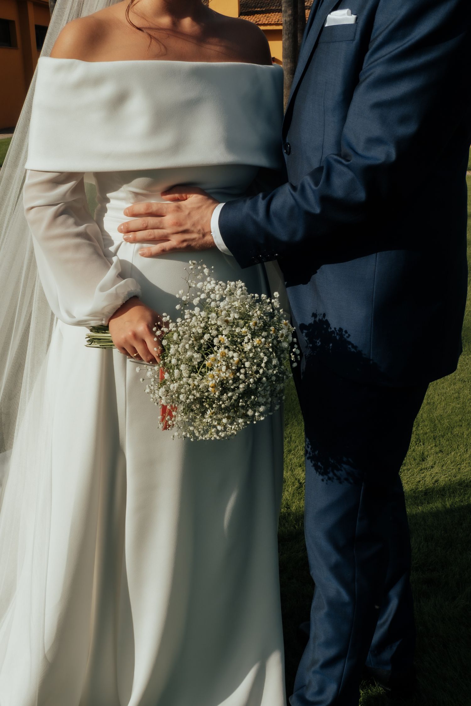 Bride holding baby's breath bouquet in off-shoulder gown as groom in navy suit embraces her waist.