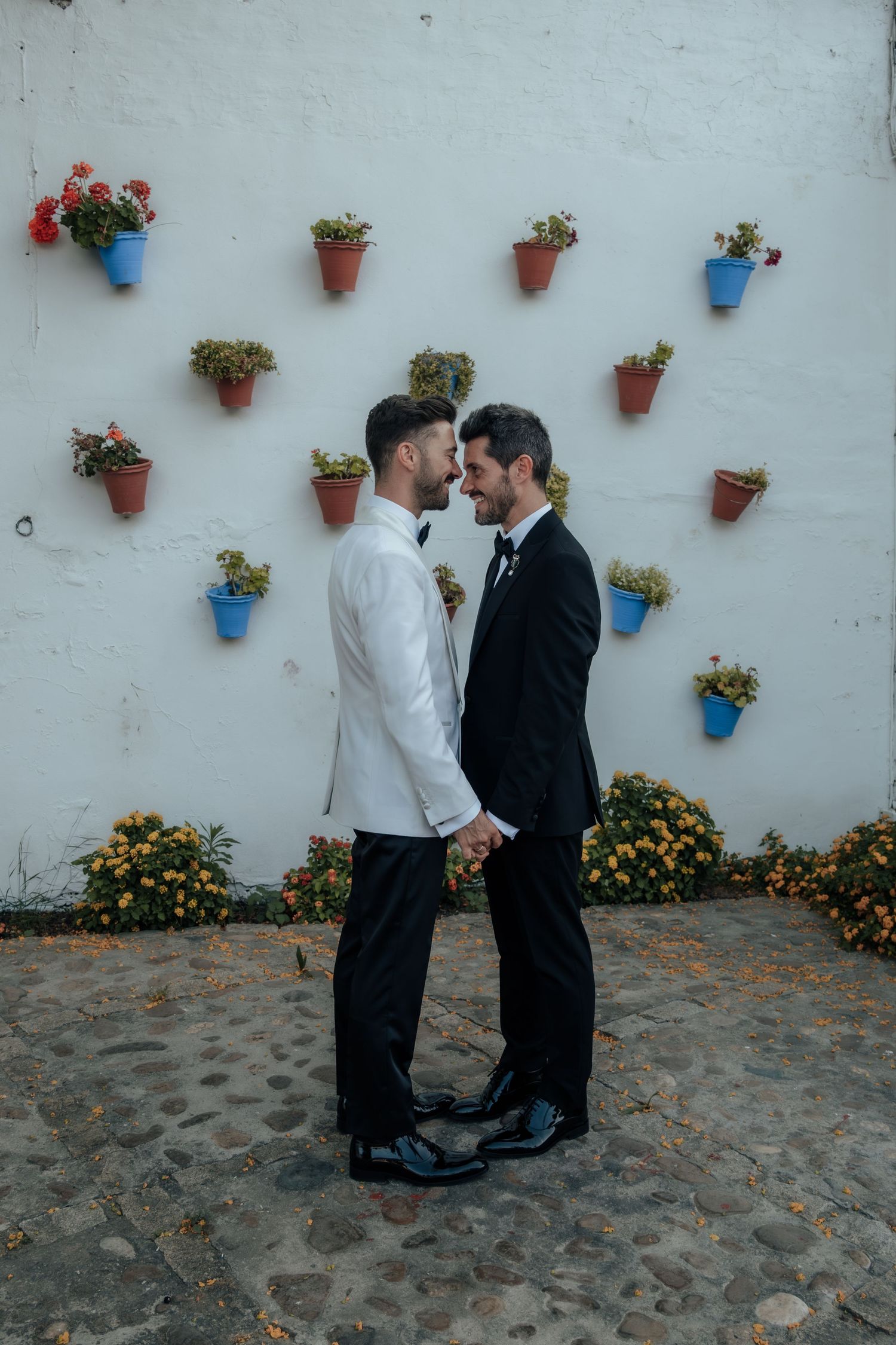 Two grooms in black and white tuxedos hold hands facing each other by floral wall.