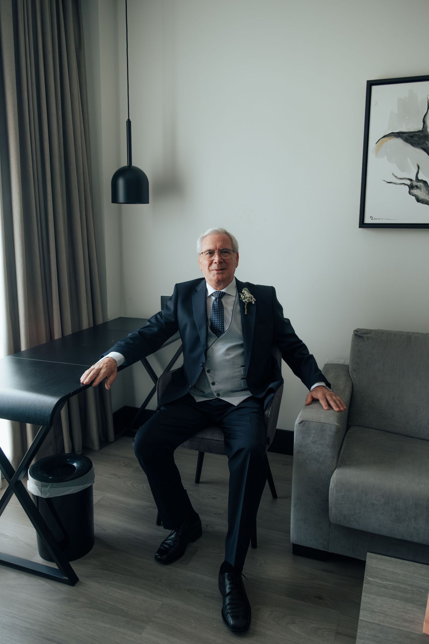 Elegant senior man in a dark suit with boutonniere sitting confidently in a modern hotel room.