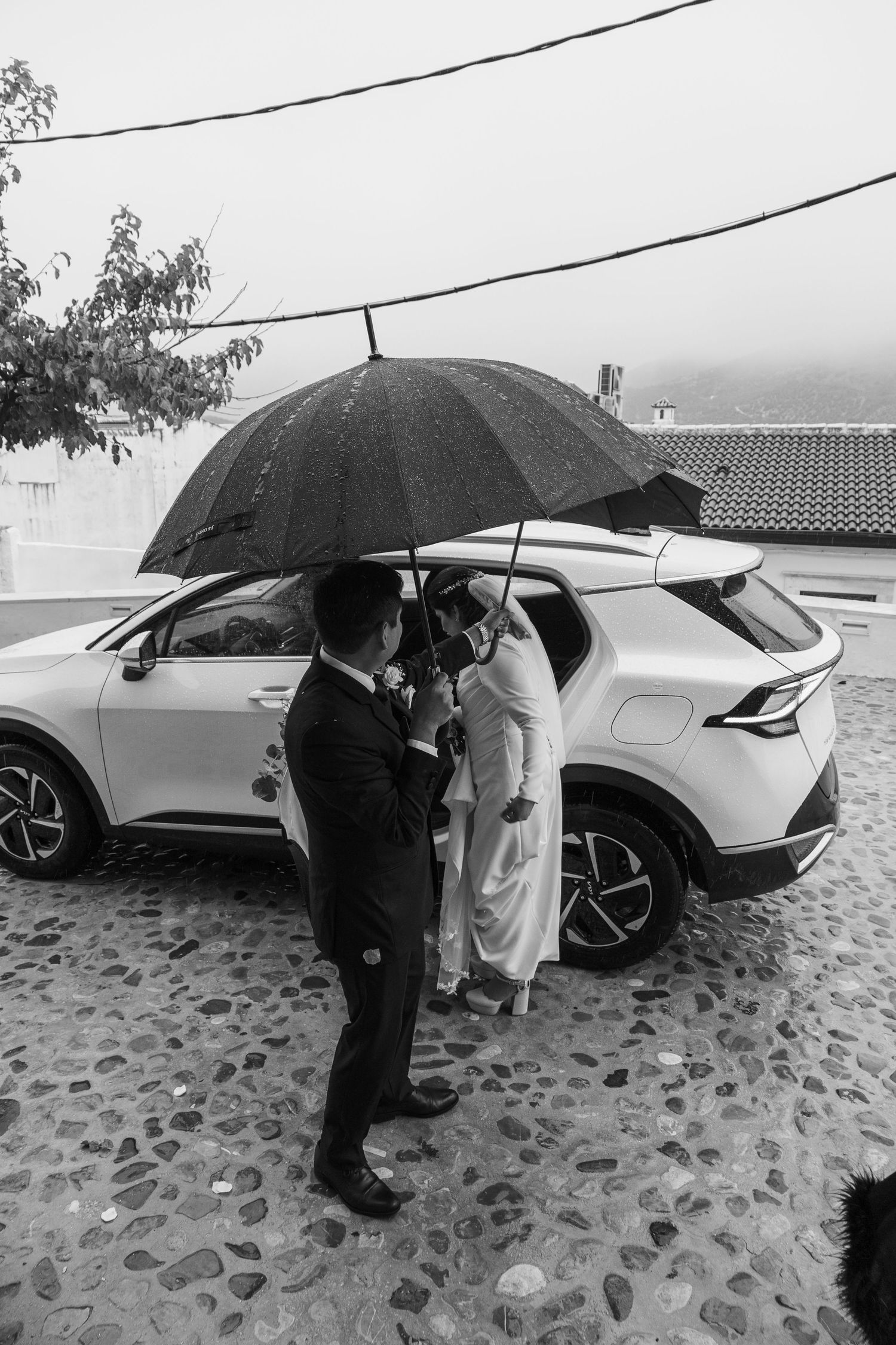 Black and white photo of wedding couple sheltering under umbrella beside white car in rain.