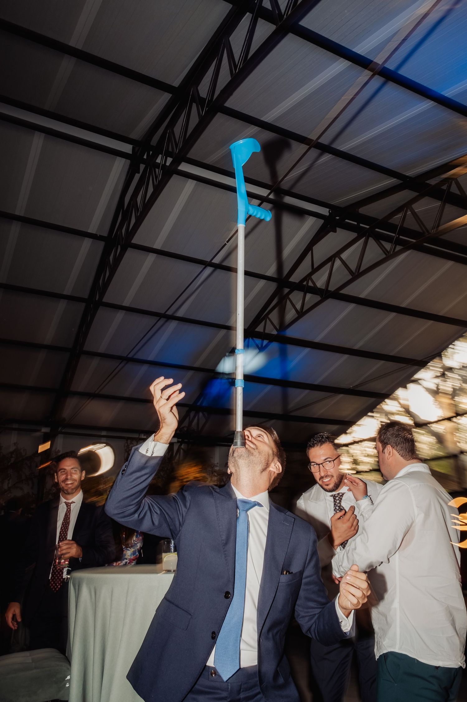 Man in blue suit playfully balancing a crutch on his chin at a lively wedding reception party.
