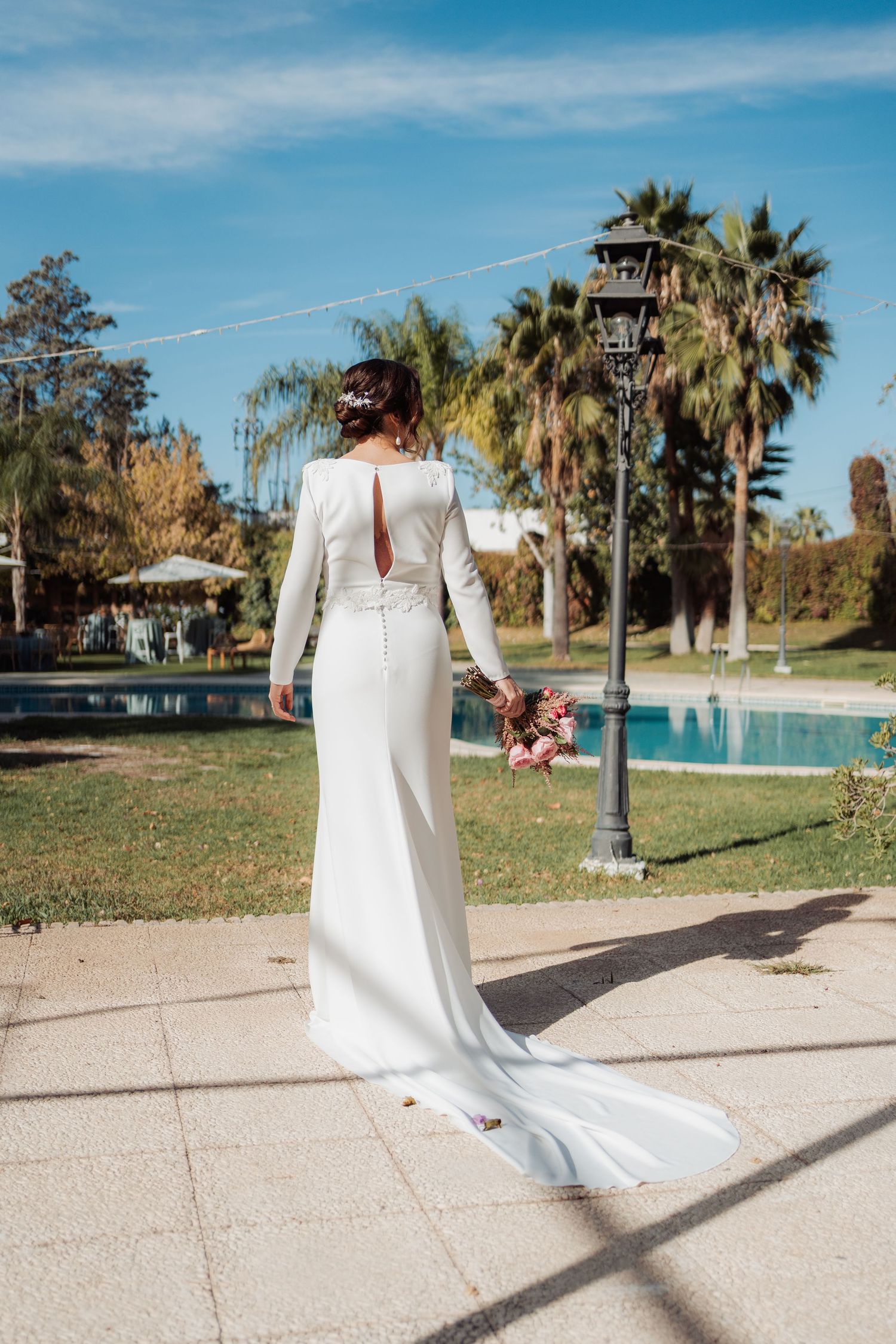 Bride in fitted white long-sleeve gown with train holding pink bouquet by poolside garden.