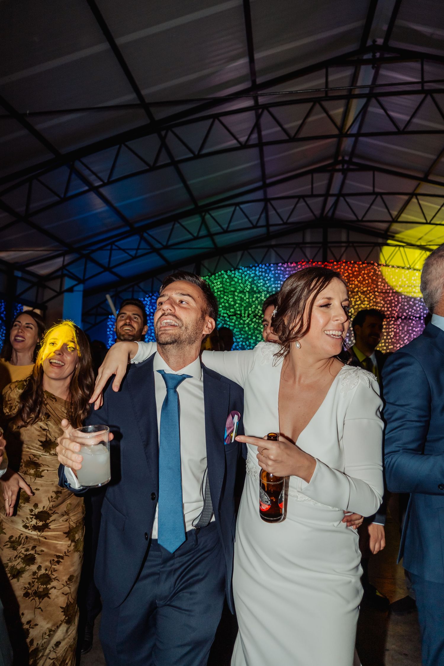 Bride and groom dancing joyfully with guests under colorful rainbow lights at wedding reception party.
