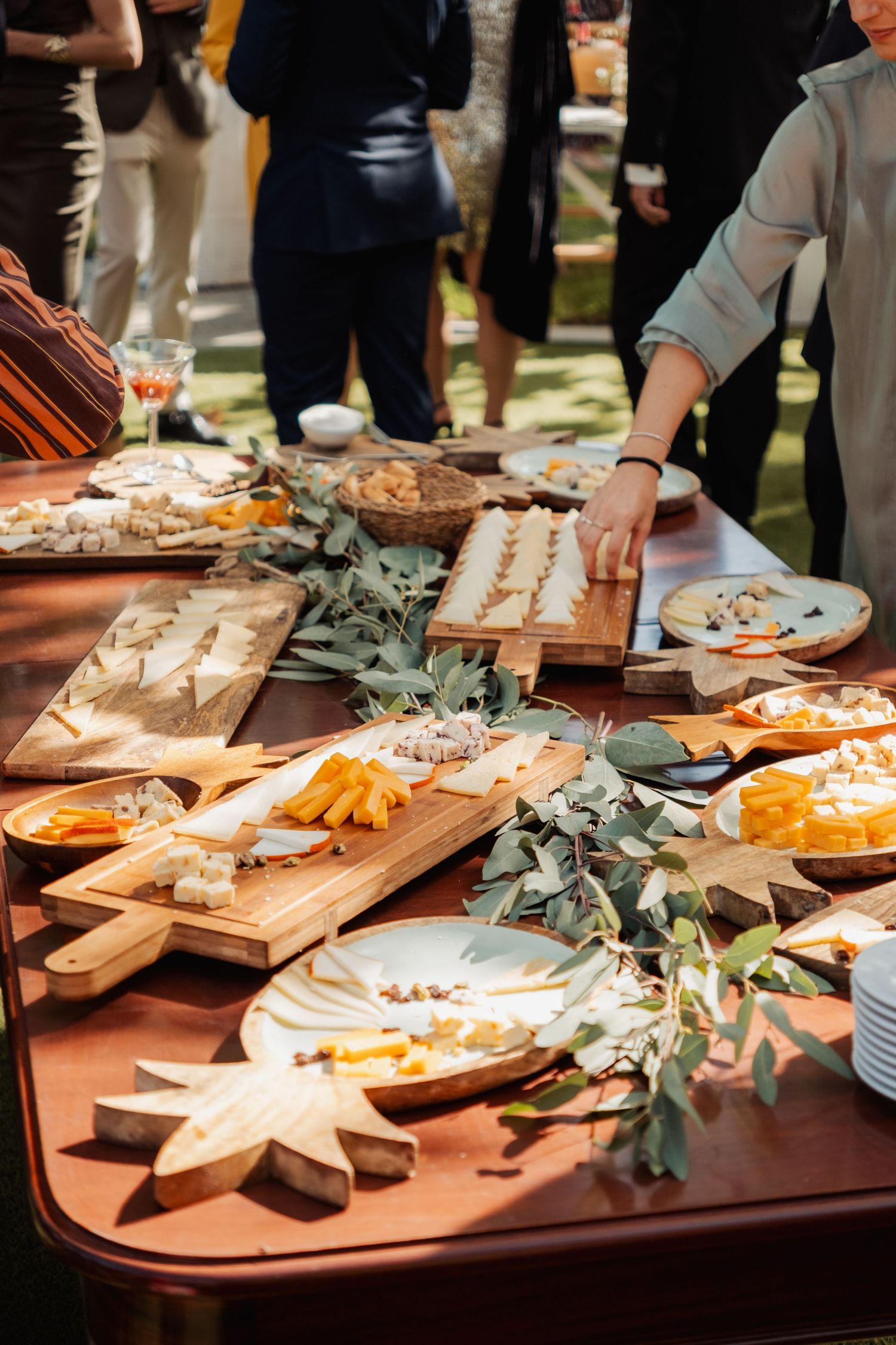 Elegant outdoor wedding catering table with cheese boards, fruit, and eucalyptus leaf decorations.