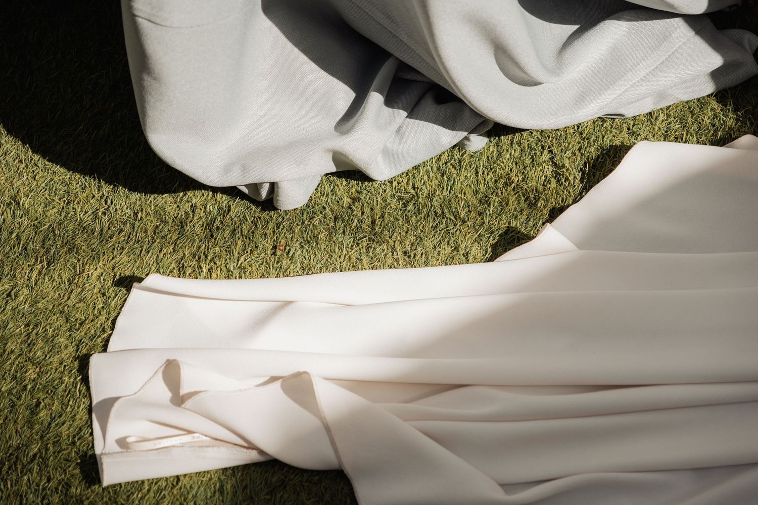 Detail shot of white wedding dress train spread elegantly on green grass in sunlight.