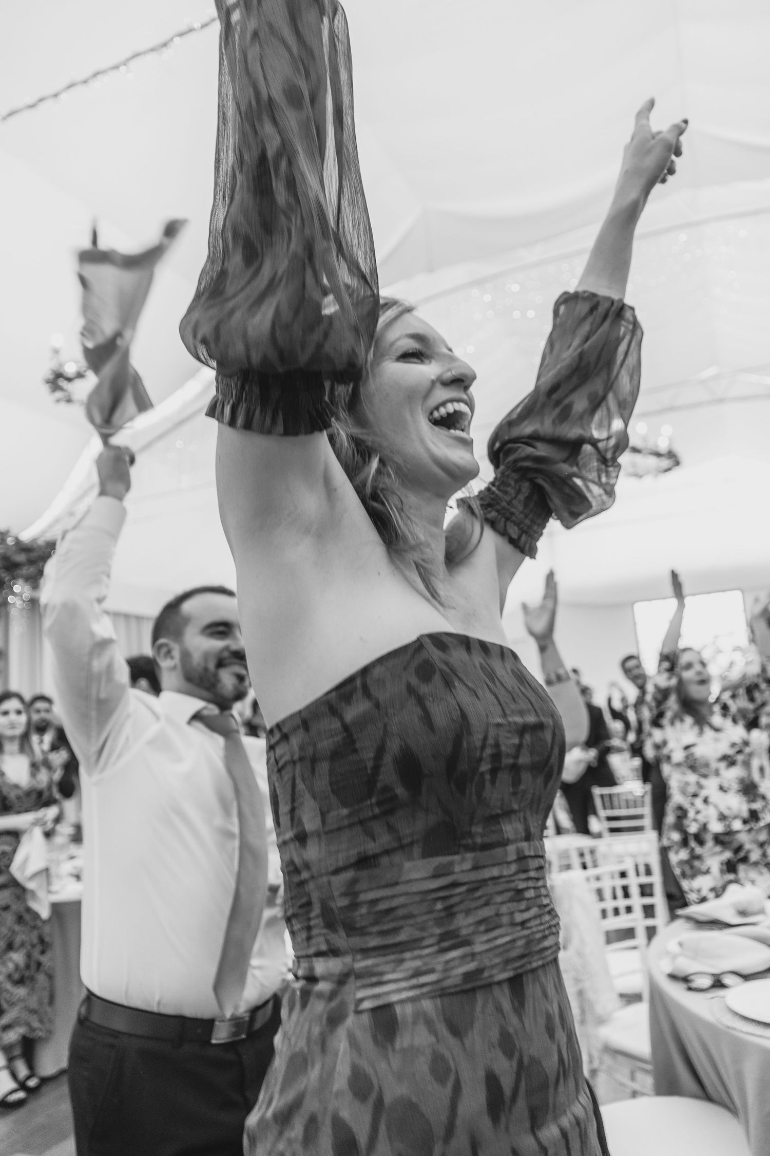 Black and white photo of laughing woman with arms raised in joy at a festive wedding reception.