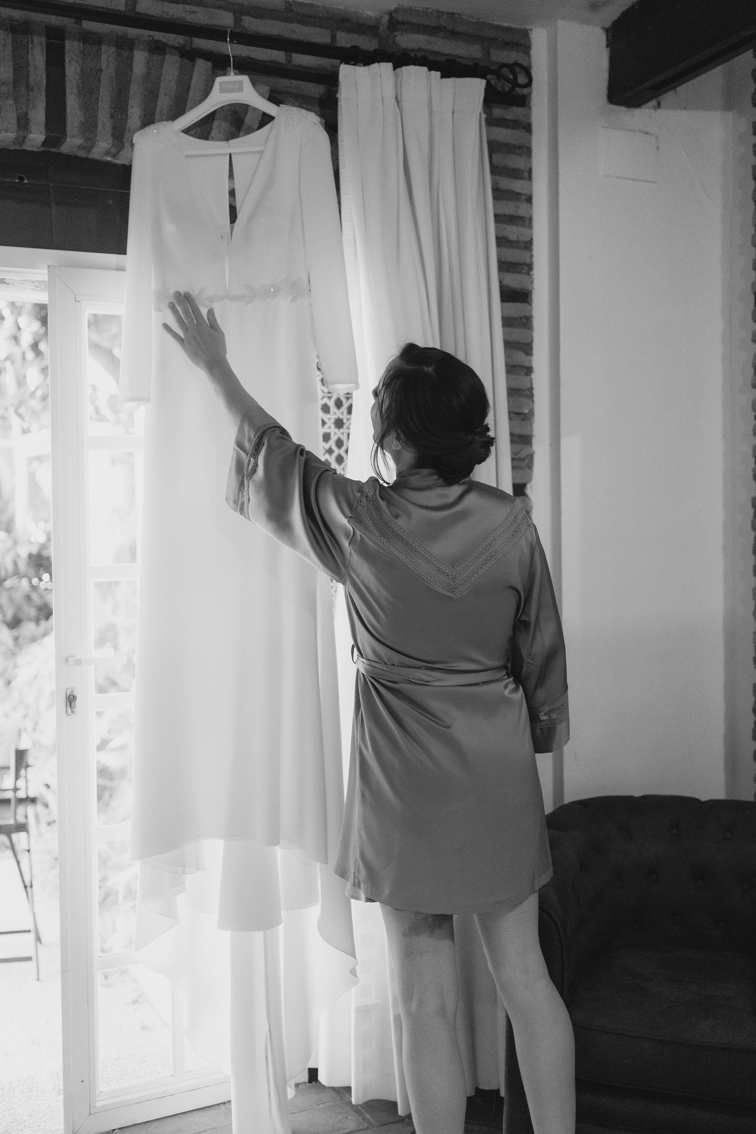 Black and white photo of bride in robe reaching for wedding dress hanging by window.