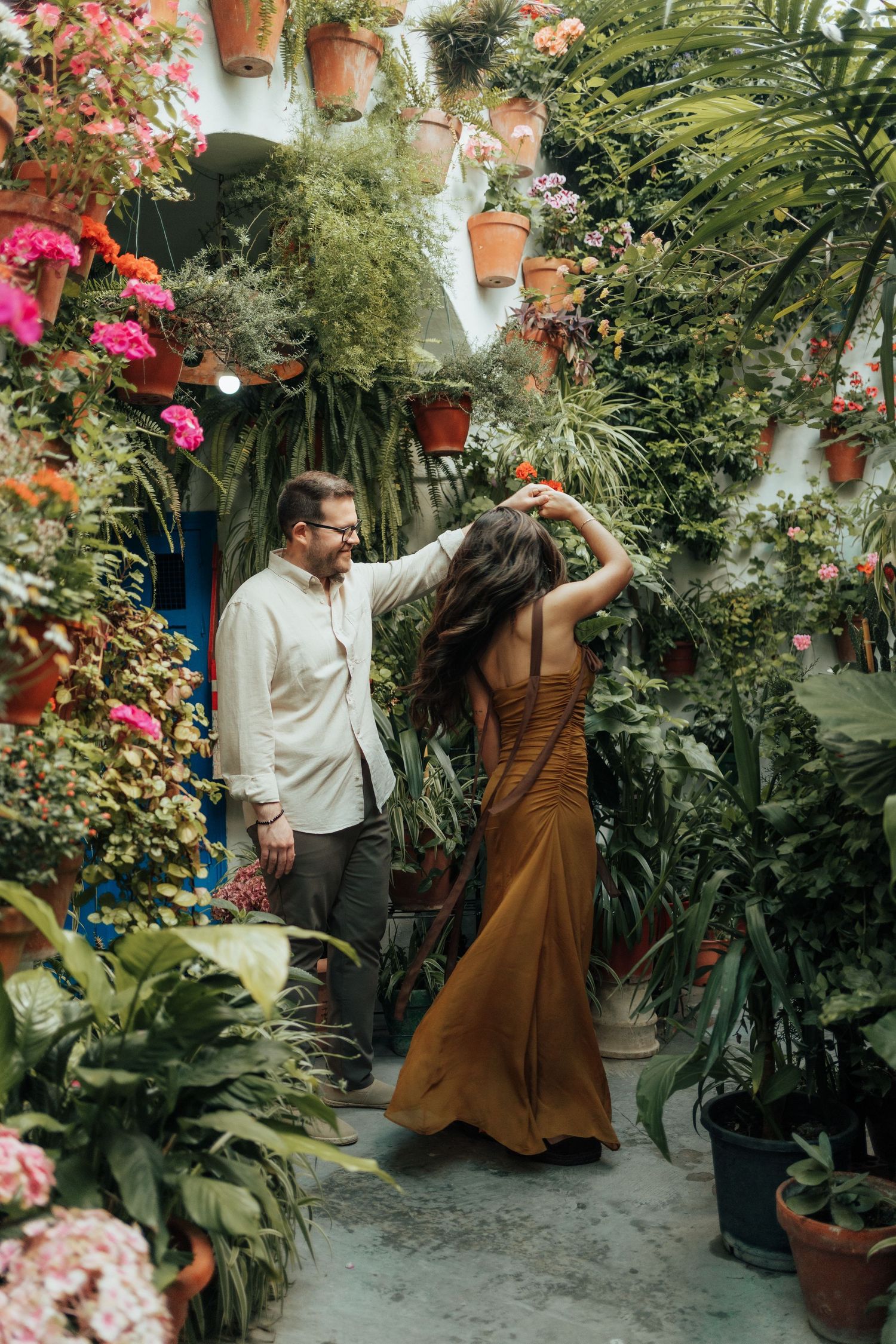 Couple dancing in a lush floral courtyard, woman in brown gown, man in white shirt, surrounded by colorful potted plants.