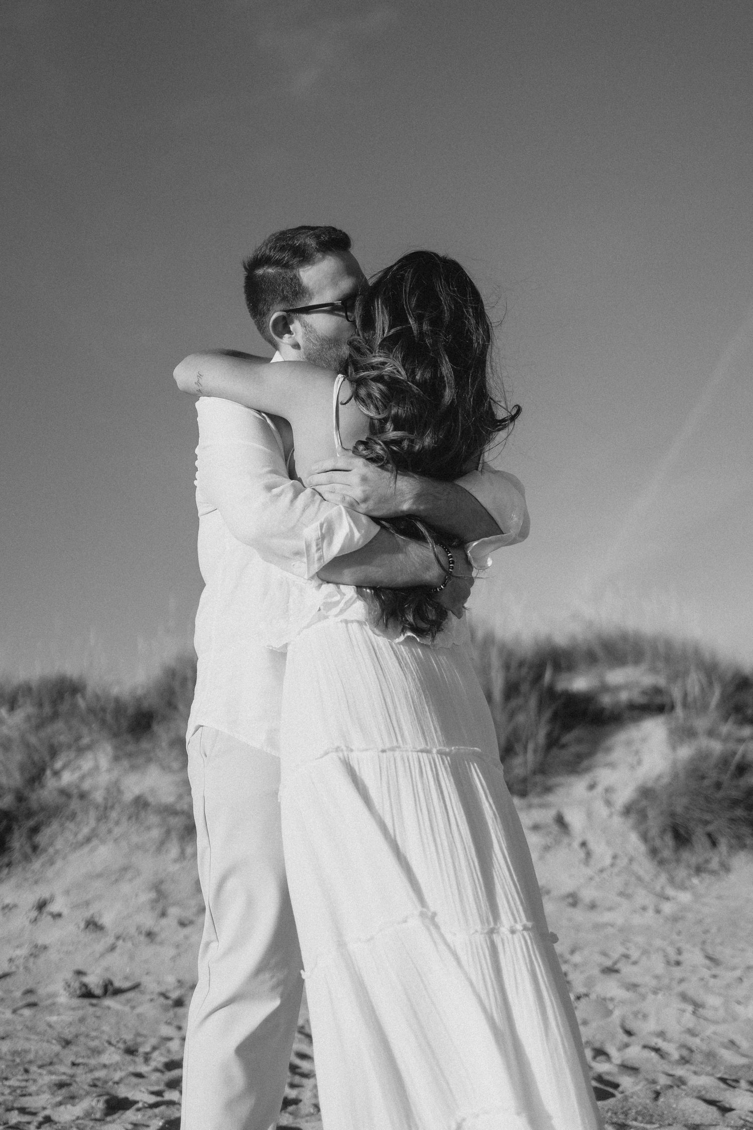 Black and white photo of a couple embracing on a sandy beach, woman in flowing white dress, dunes in background.