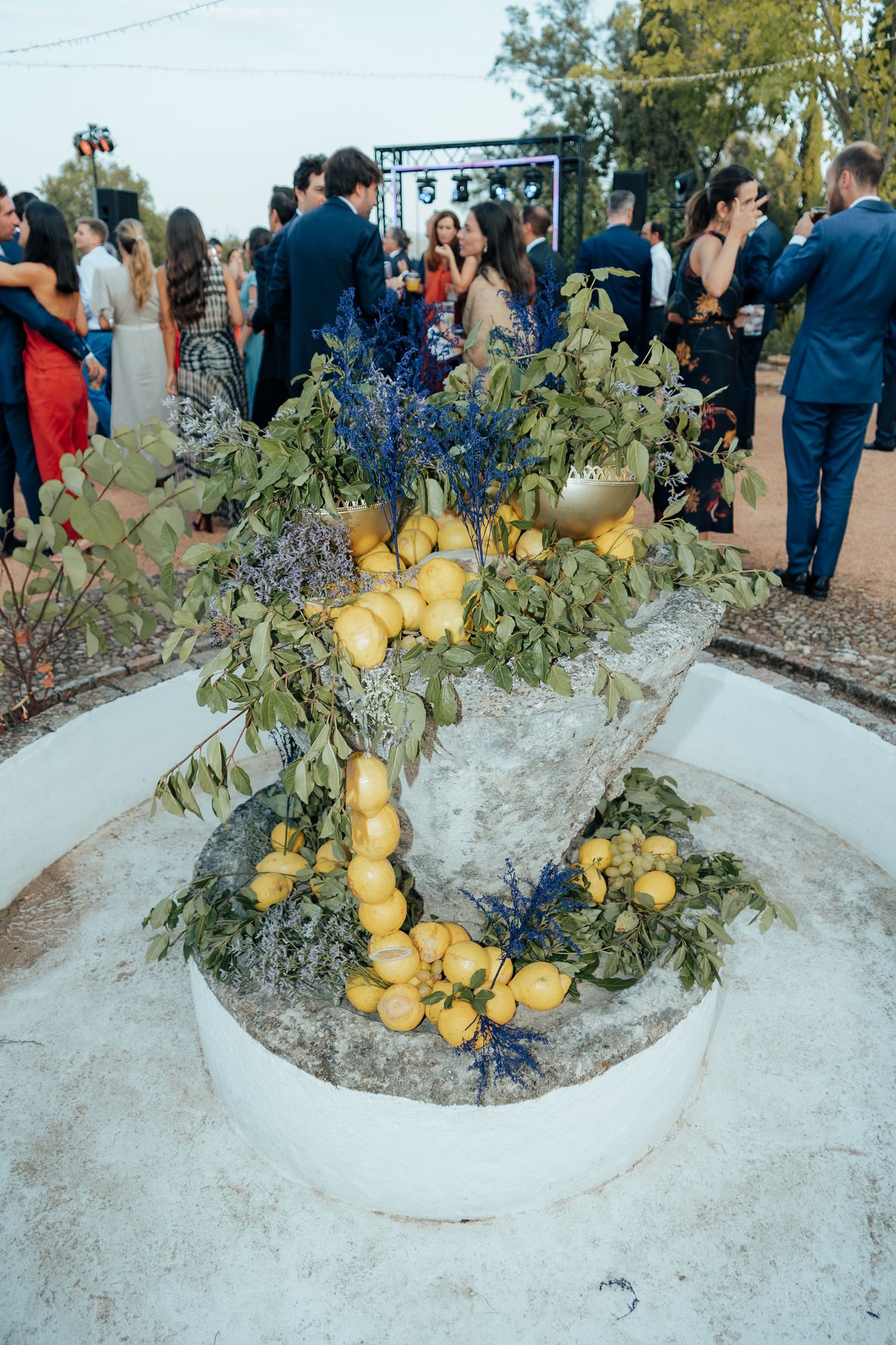 Decorative stone fountain adorned with lemons, green leaves, and blue flowers at wedding venue.