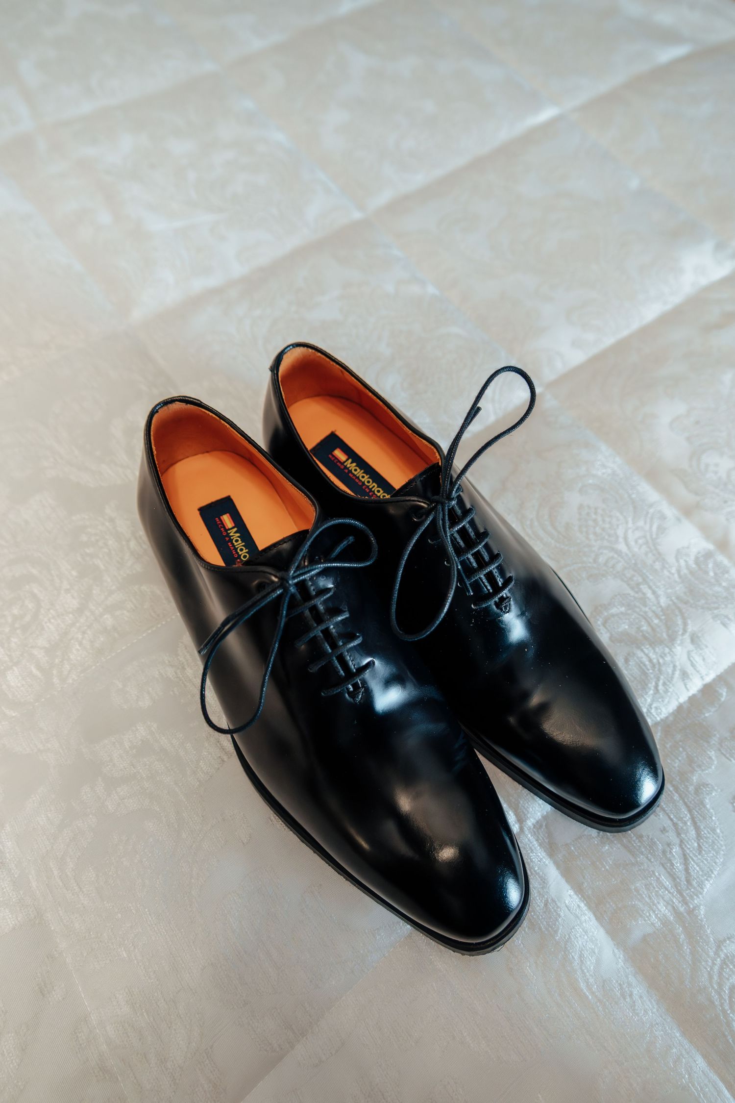Elegant black Oxford dress shoes with orange insoles placed on white embroidered fabric surface.
