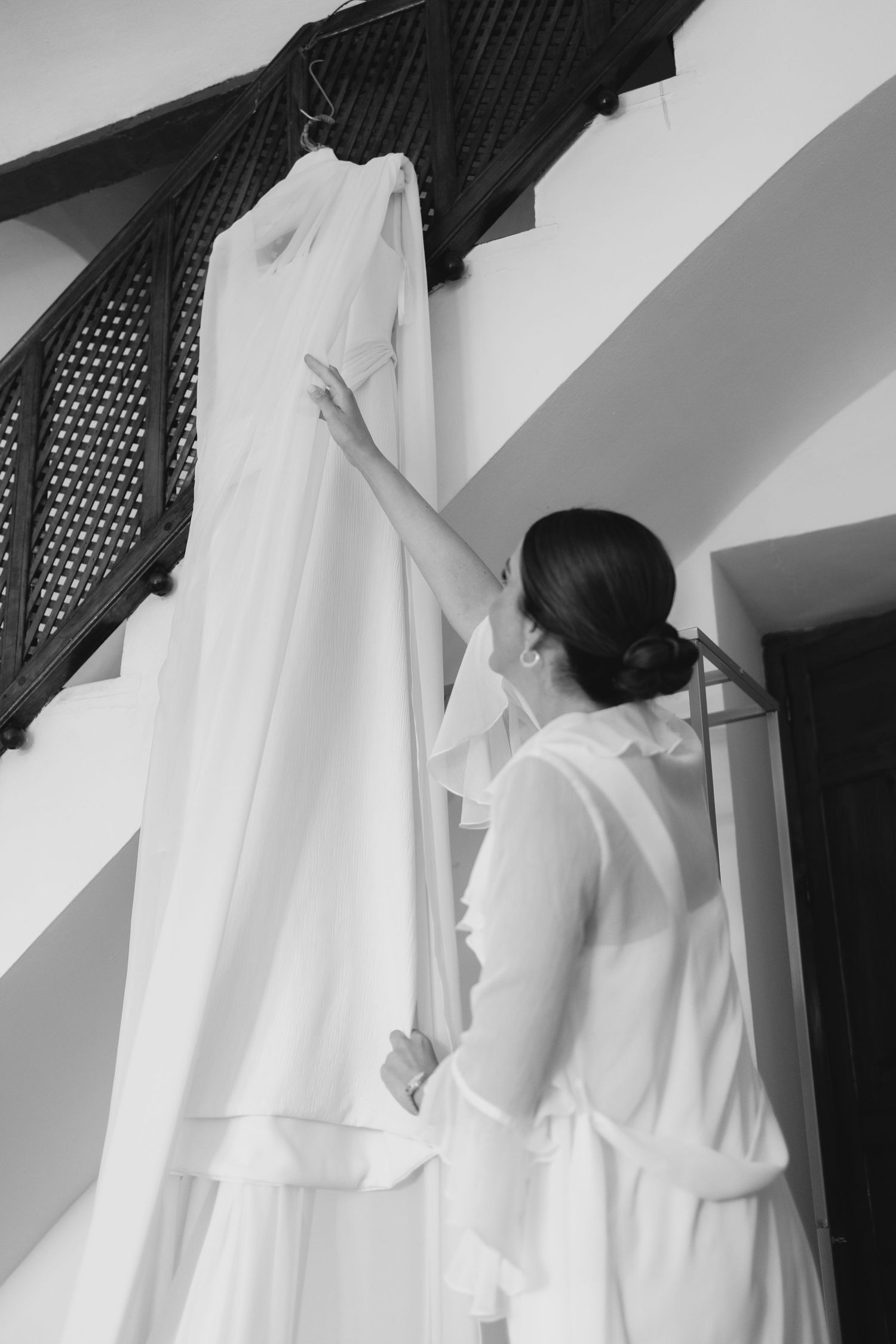 Black and white photo of bride reaching for her wedding dress hanging on staircase.