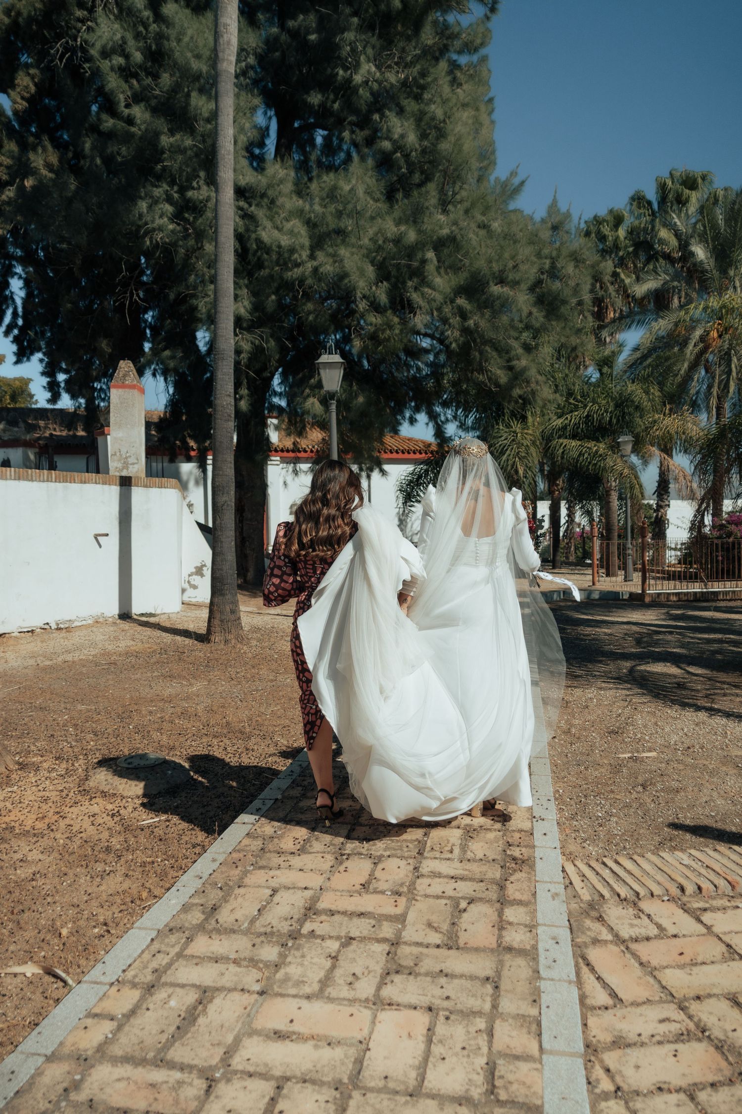 Wedding helper adjusts bride's flowing white gown and veil on a brick pathway lined with tall trees.