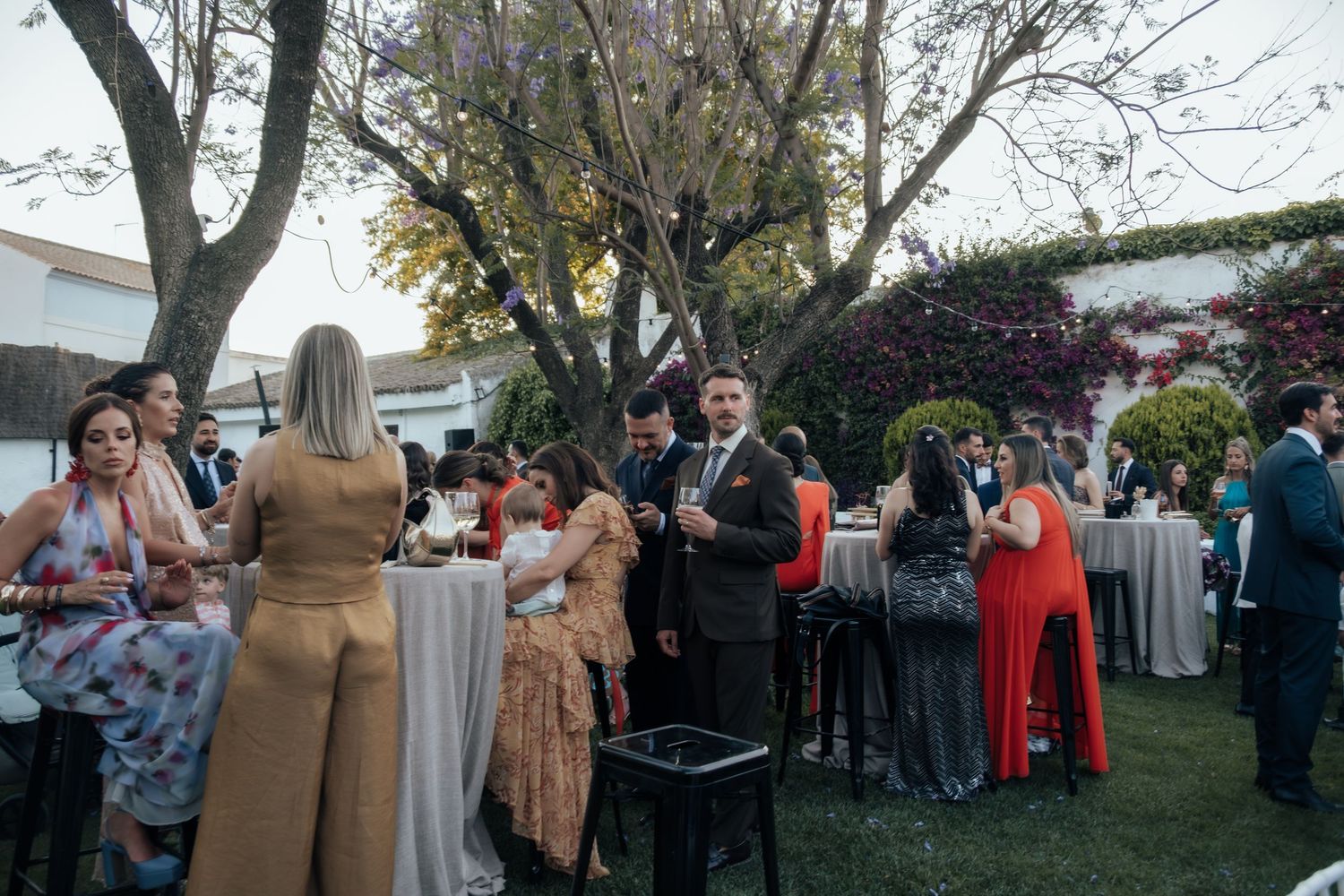 Wedding guests mingling outdoors under trees at elegant garden cocktail reception party.