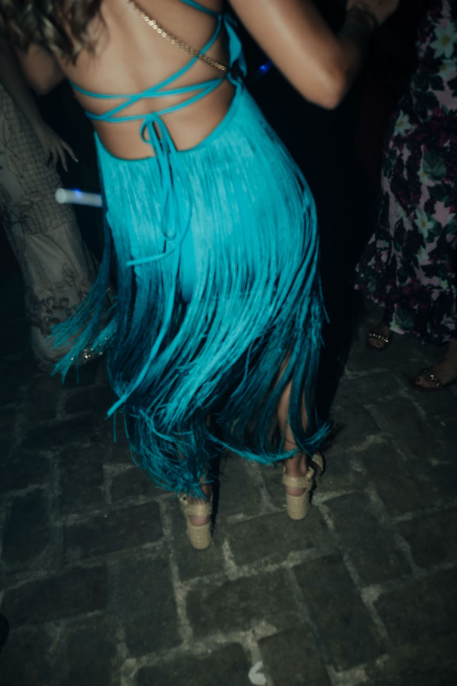 Woman in turquoise fringe backless dress dancing on cobblestone floor at an evening party event.