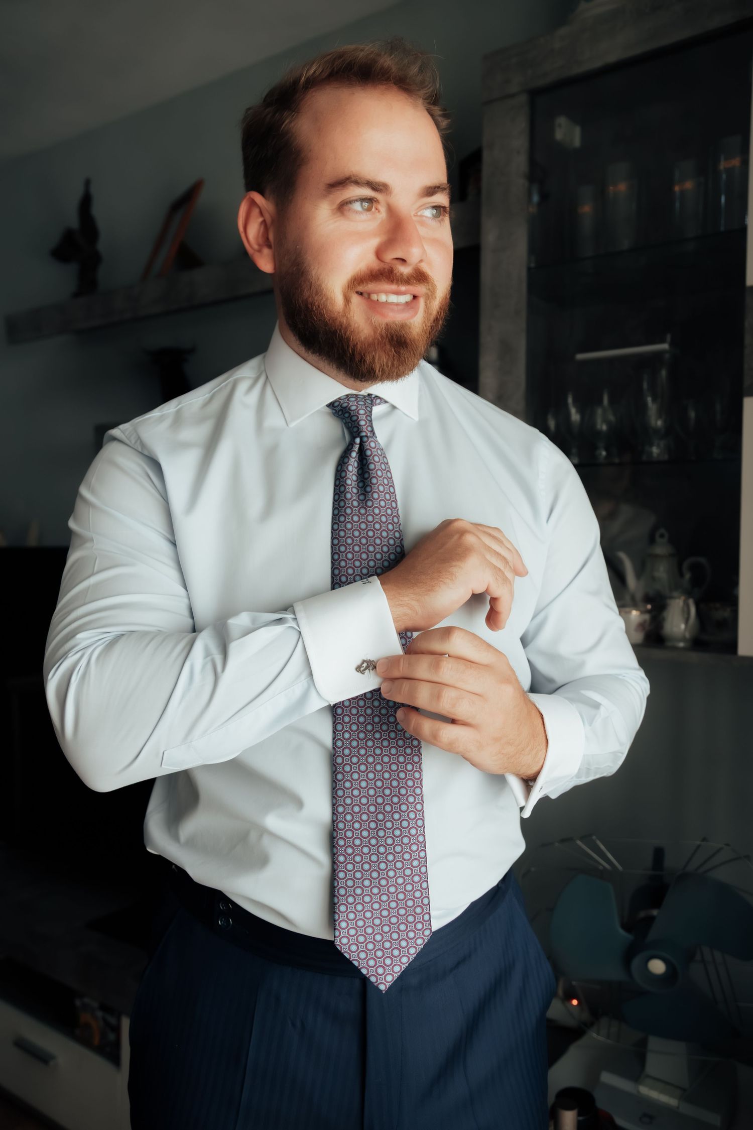 Smiling bearded groom in white shirt and patterned tie fastening cufflinks while getting ready.