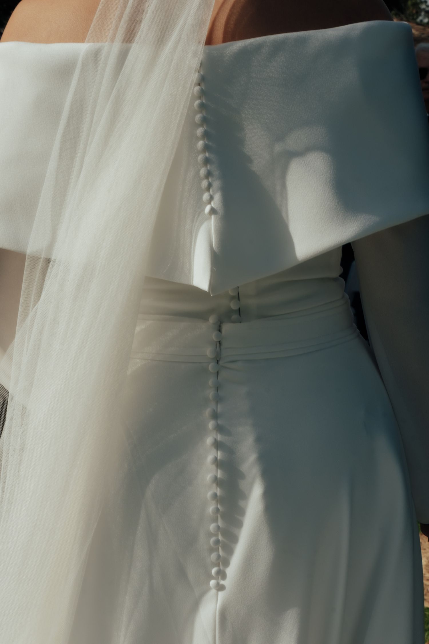 Close-up detail of bridal gown showing button back closure and flowing tulle veil in sunlight.