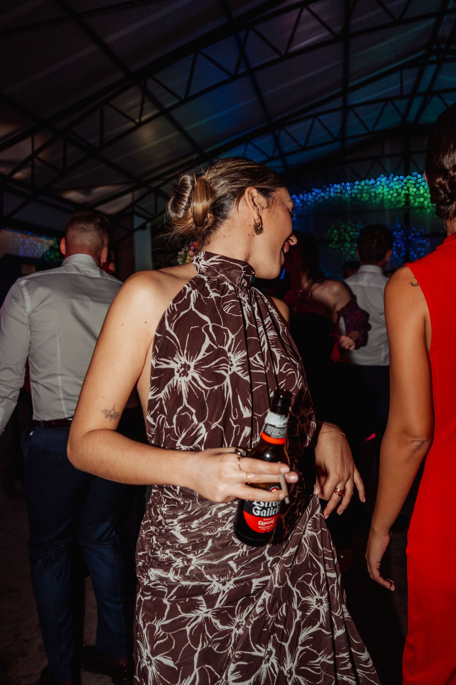 Woman in brown floral halter dress dancing with beer bottle at vibrant wedding reception party.