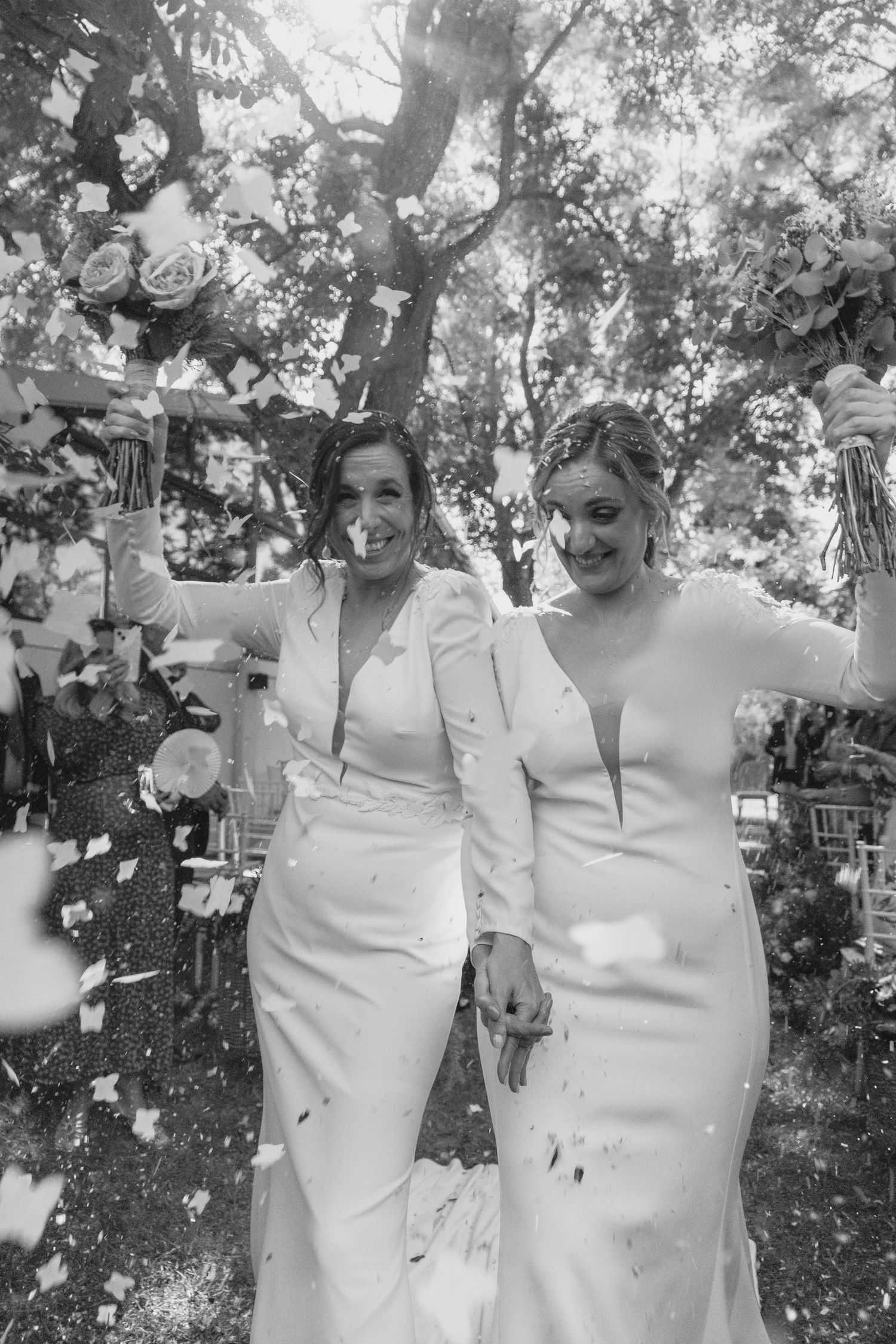 Black and white photo of two brides celebrating with raised bouquets amid falling confetti at wedding.