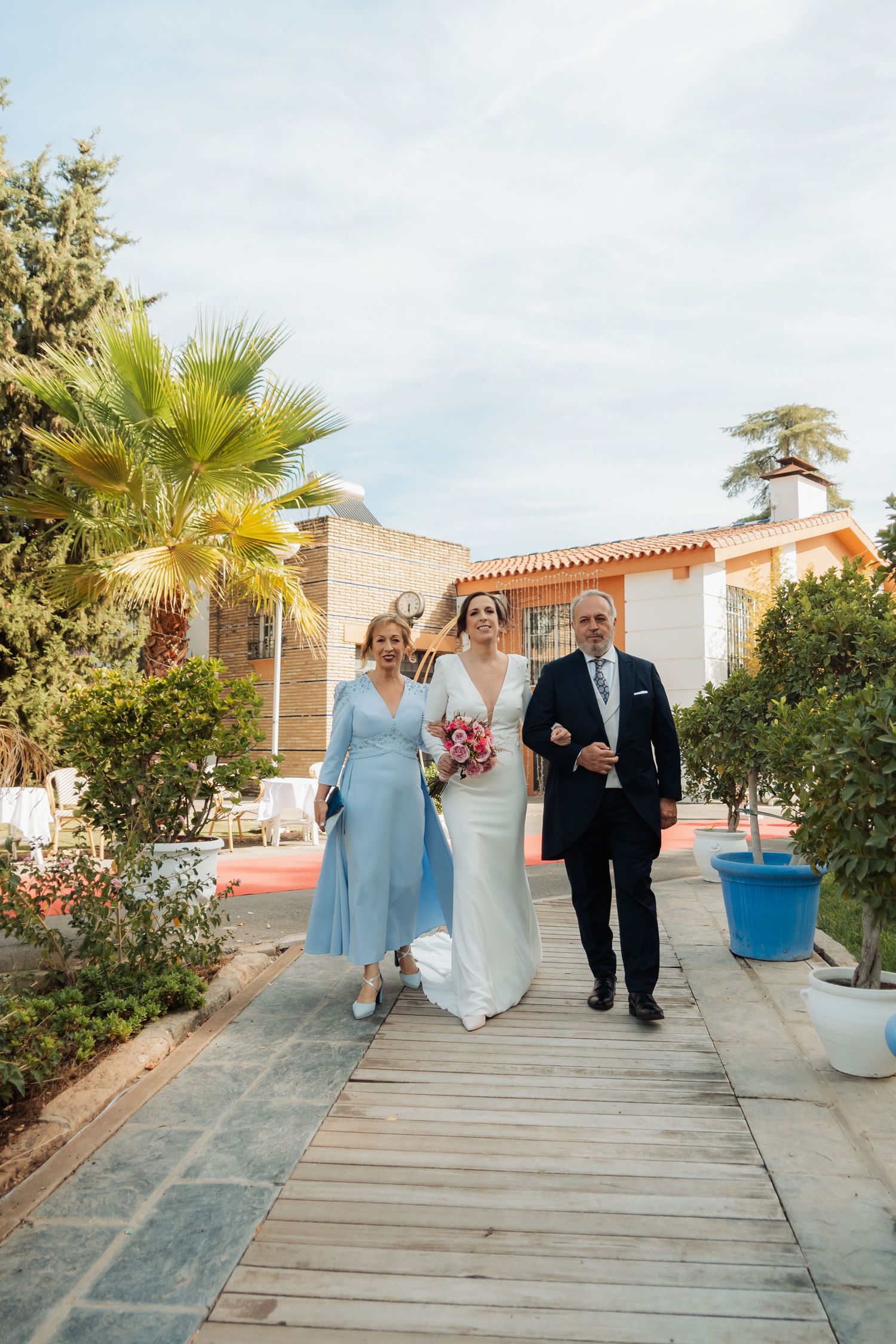 Bride in white dress walks with parents along wooden pathway at wedding venue.