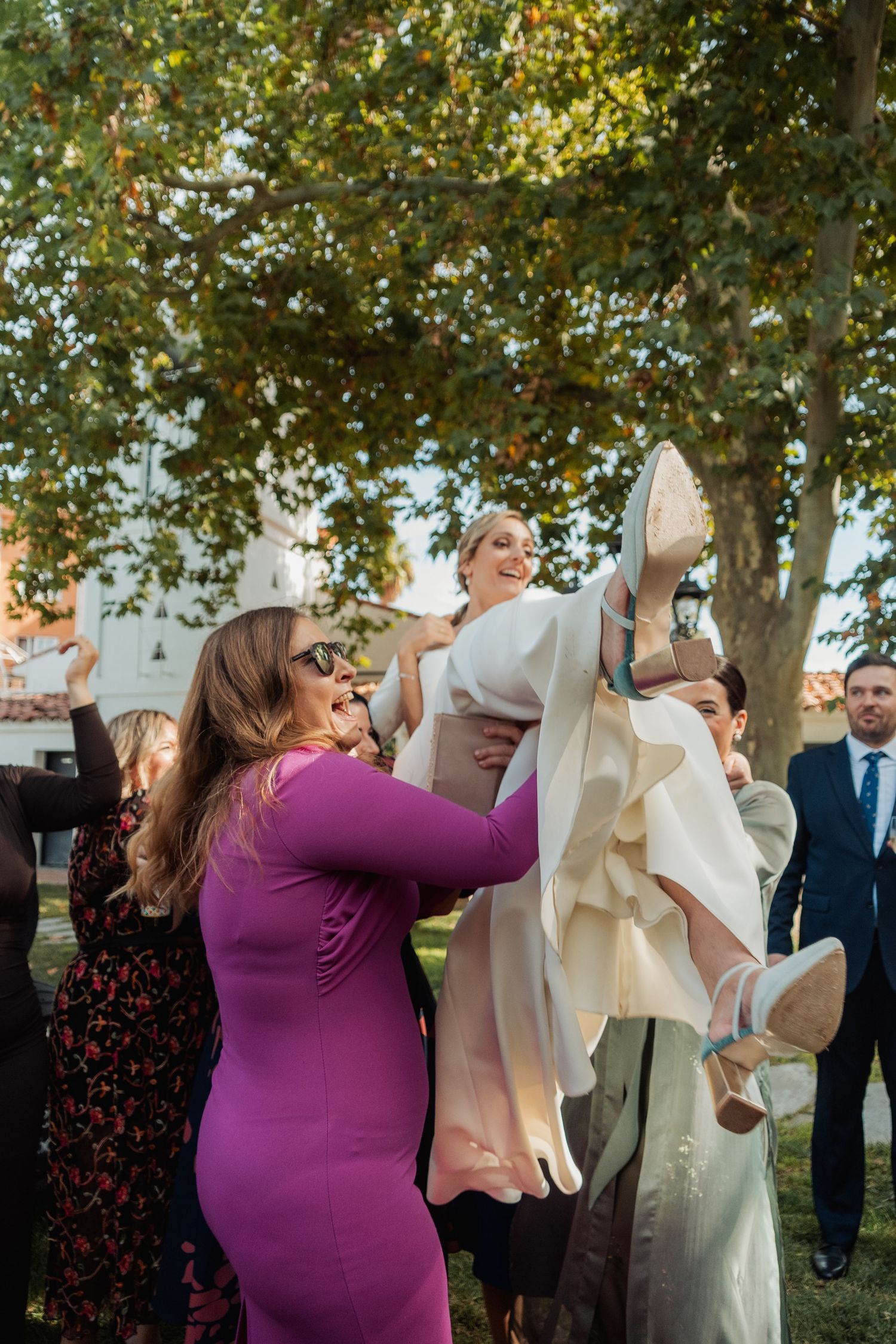 Joyful bride lifted by wedding guests outdoors while holding up her shoe in celebration.