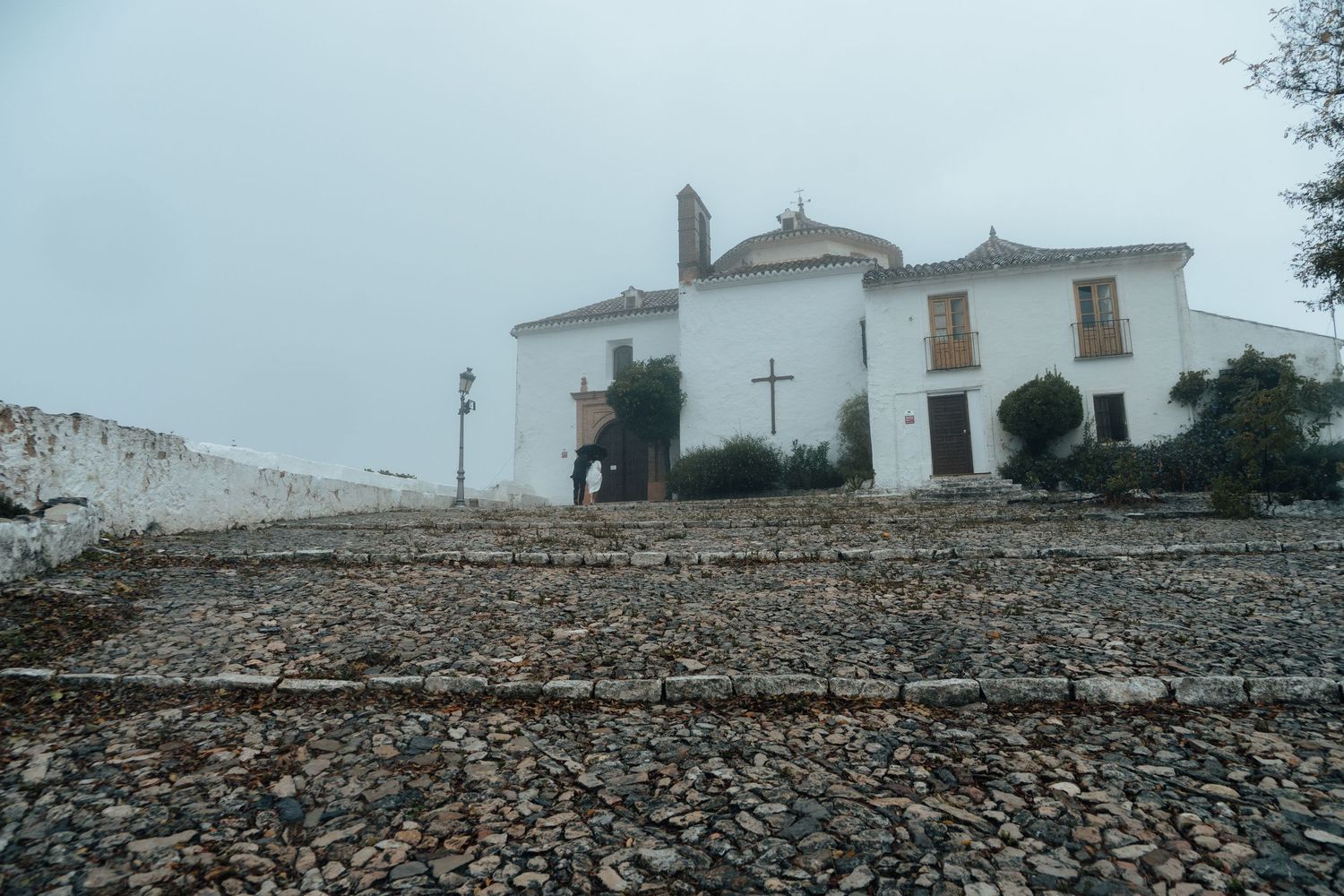 Misty view of white Spanish chapel with cross on foggy rainy wedding day.