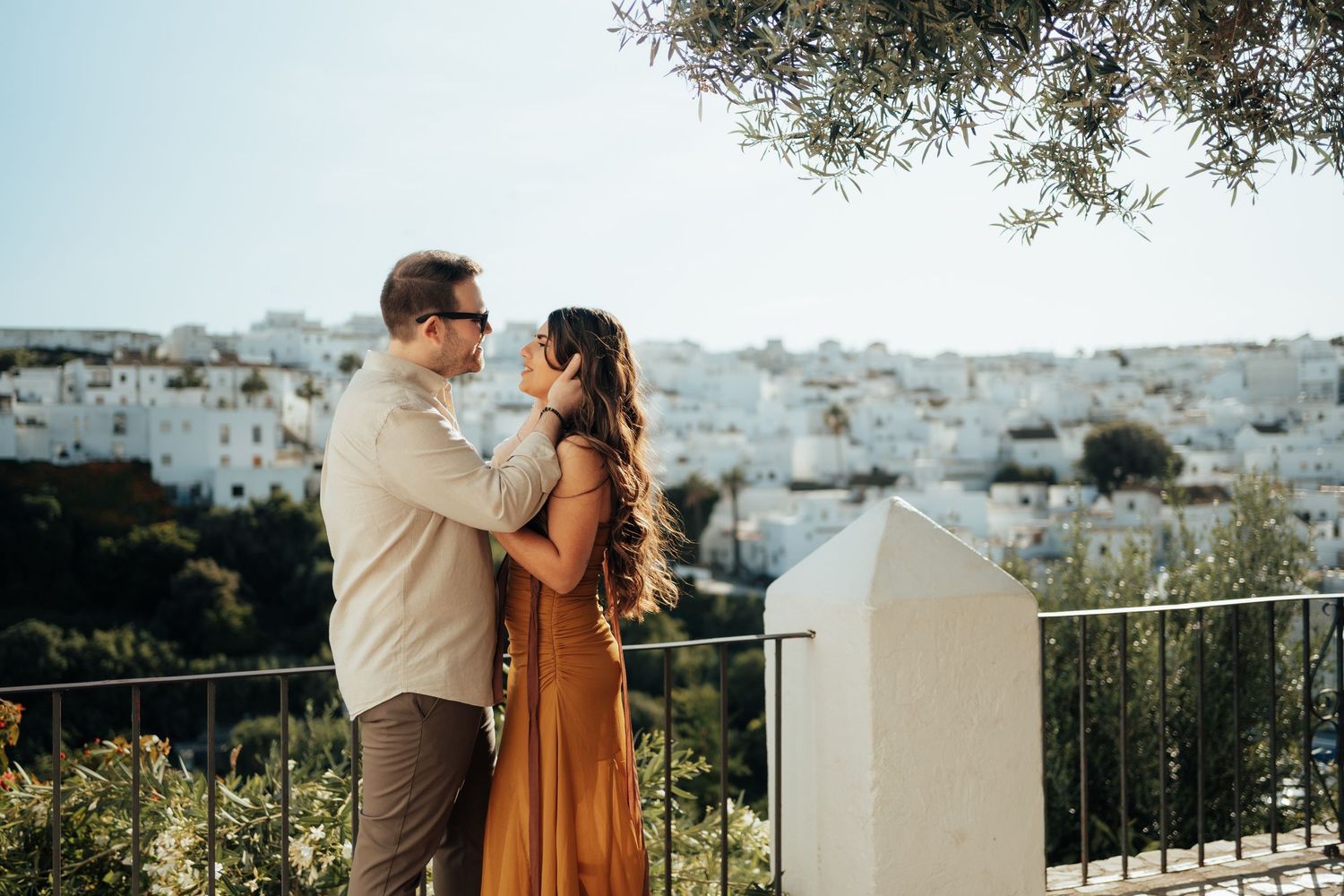 Couple embracing on a terrace overlooking a white-washed Mediterranean hillside town on a sunny day.