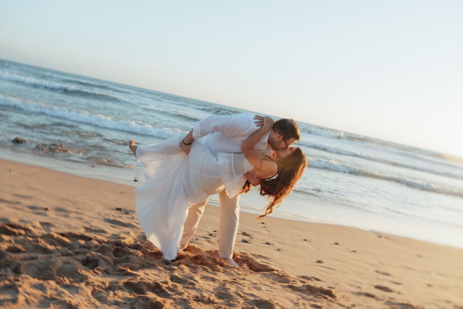 Bride and groom sharing a romantic dip kiss on a sandy beach at sunset in wedding attire.