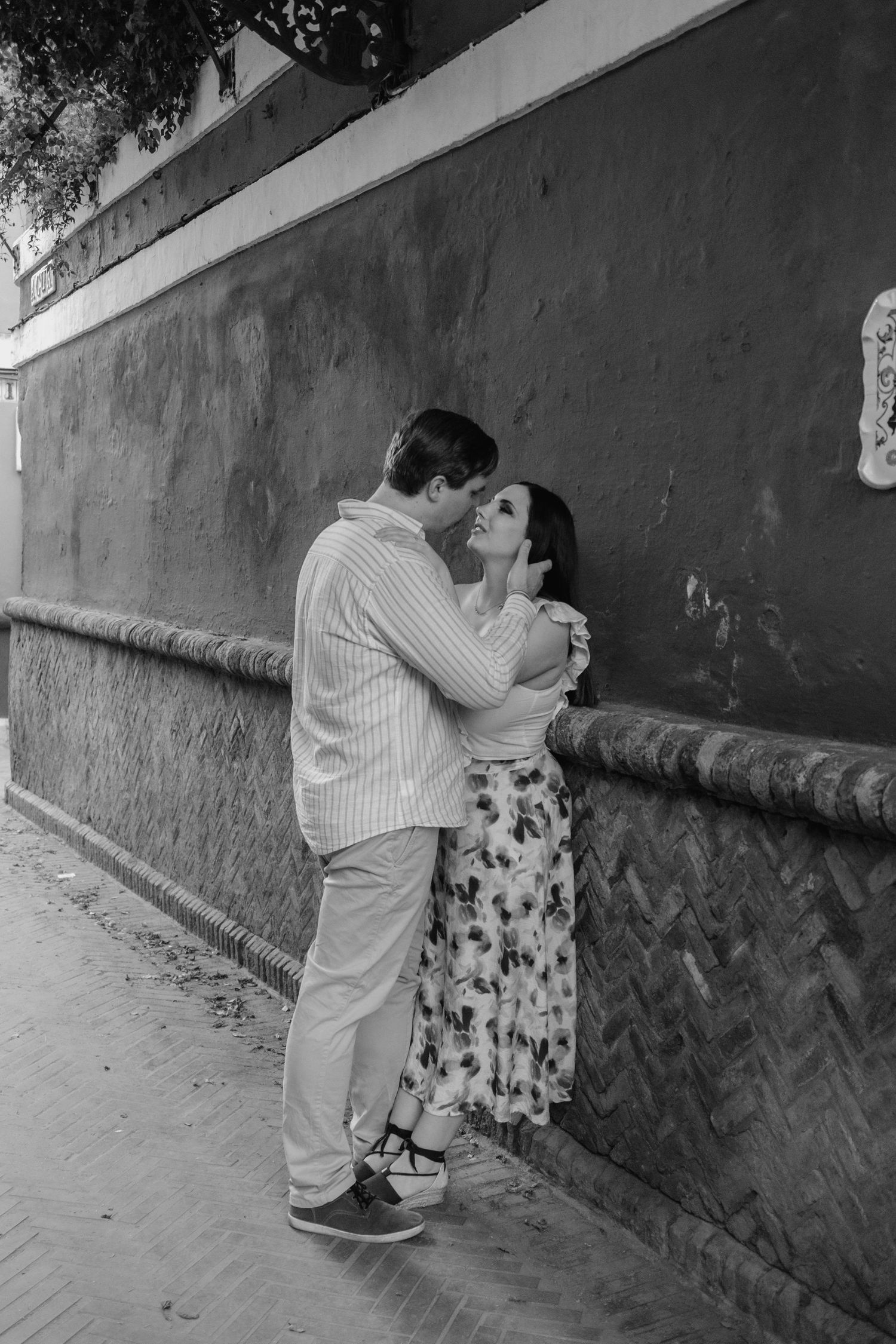 Black and white photo of a romantic couple kissing against a textured red wall on a cobblestone street.