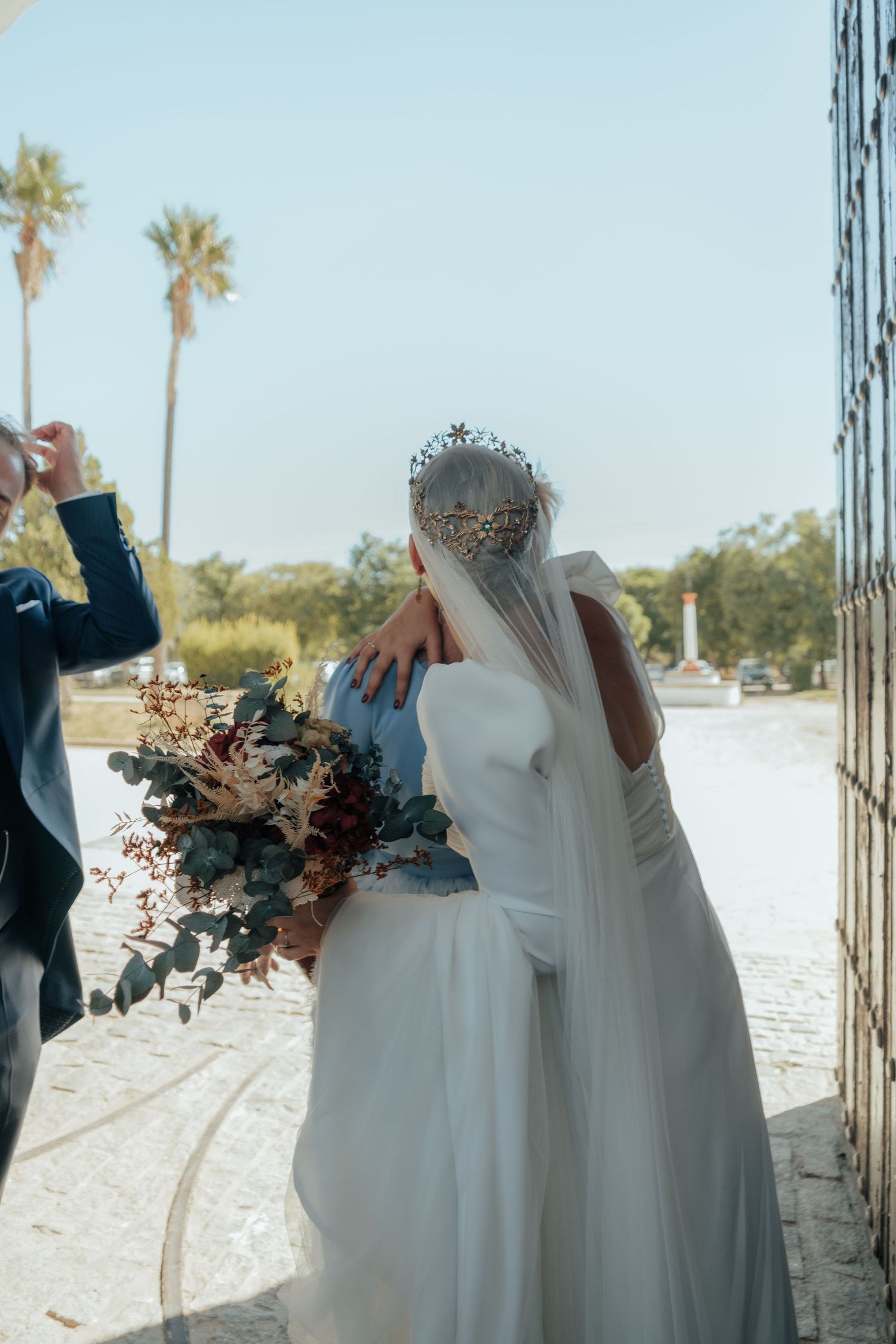 Bride in white gown with ornate crown and veil embraces groom holding an autumn floral bouquet.
