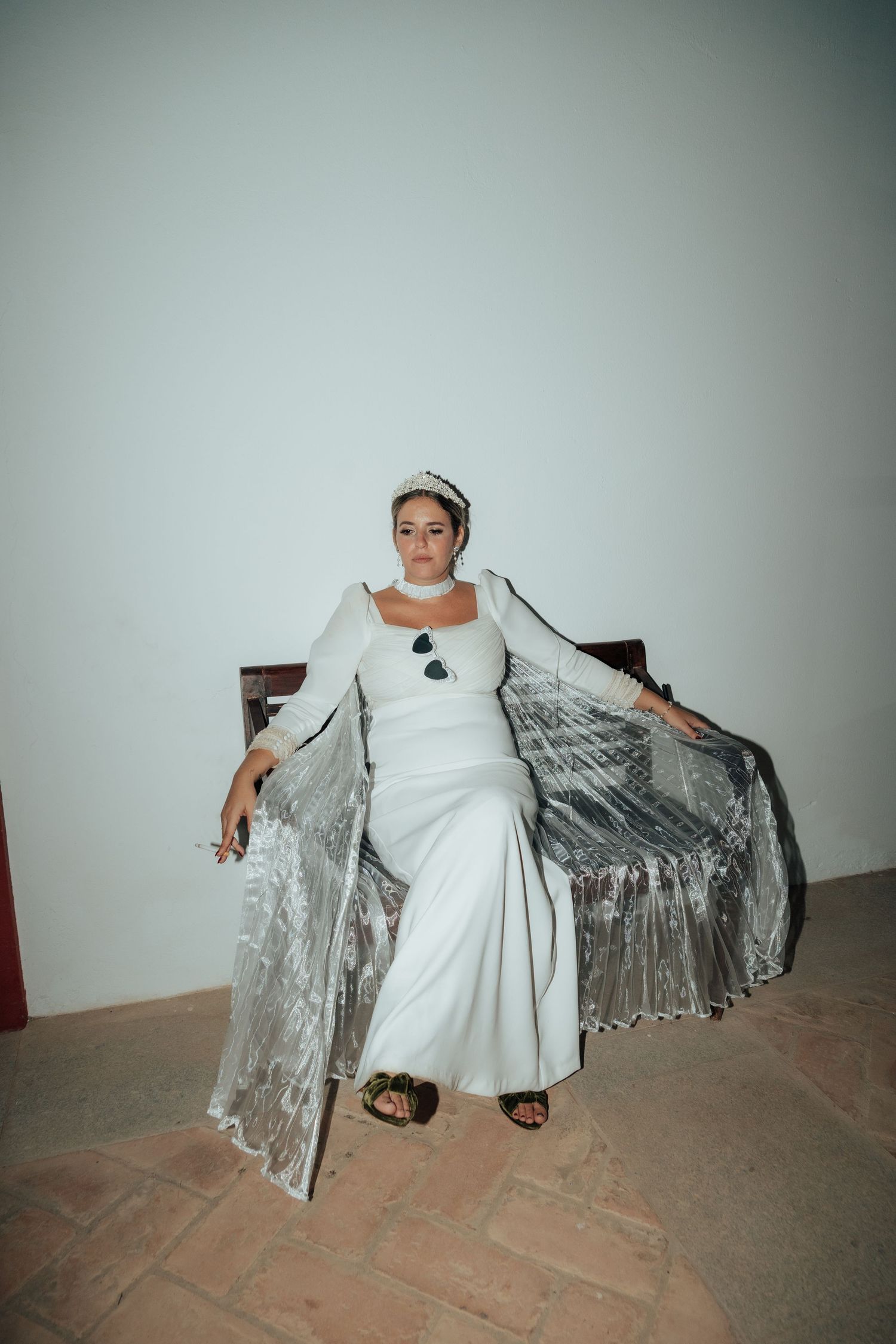 Bride in white gown and tiara seated regally on a chair against a white wall.