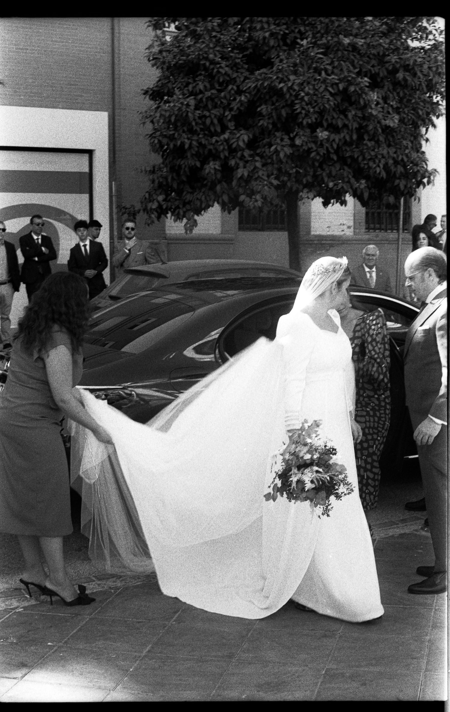 Black and white photo of bride in white gown with long veil entering a car outside a building.