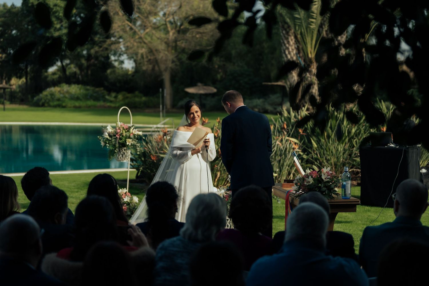 Bride and groom exchanging vows at outdoor wedding ceremony beside a pool surrounded by guests.
