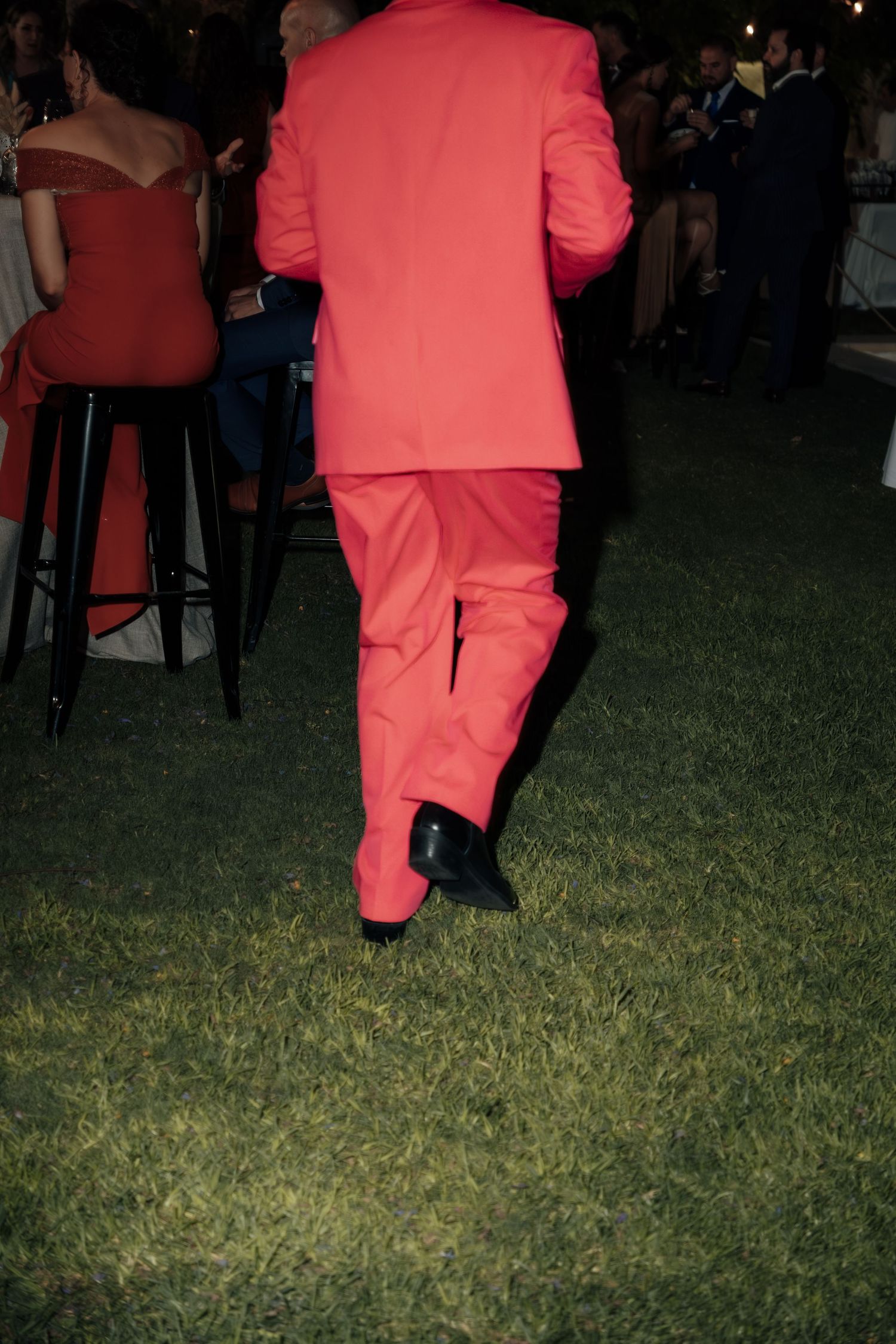 Man in bold red suit walking across grass lawn at an outdoor evening social event.