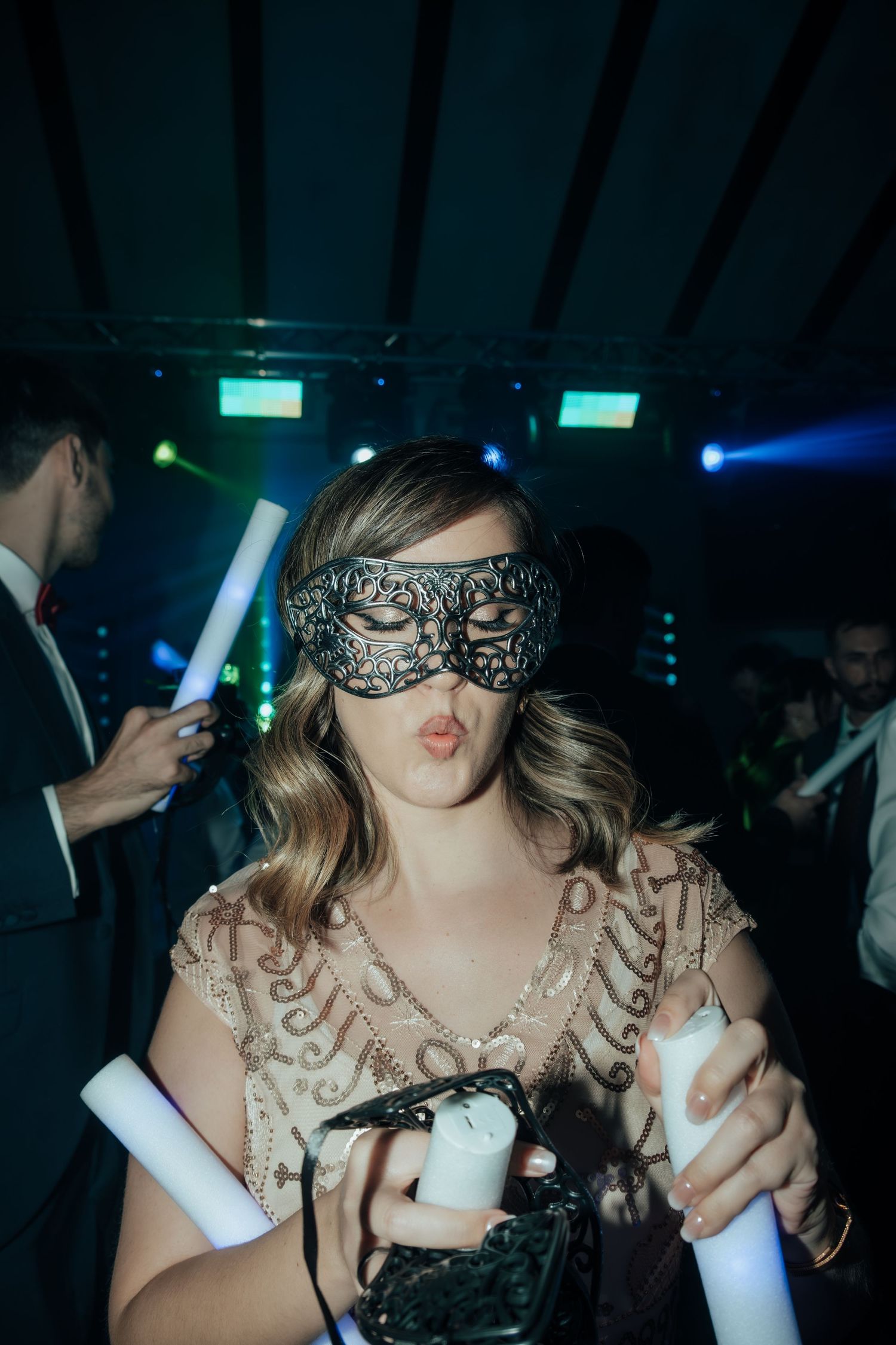 Woman in sequined dress wearing black lace masquerade mask holding glow sticks at wedding party.