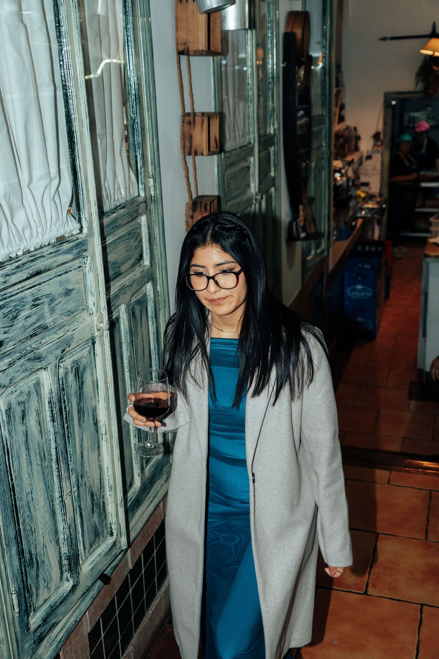 Elegant woman with glasses holding a glass of red wine near vintage wooden shutters indoors.