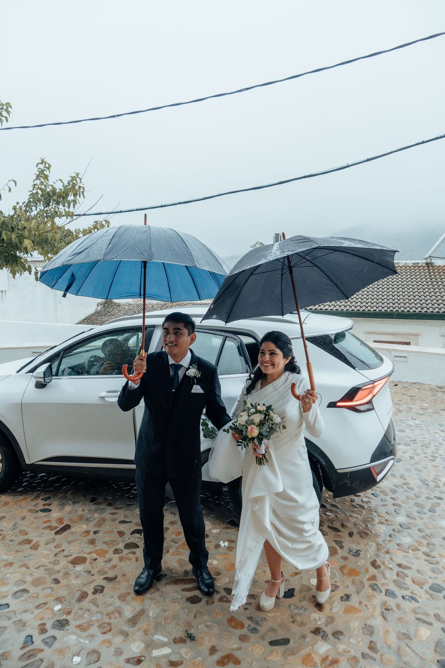 Bride and groom holding umbrellas in the rain on their wedding day beside a white car.