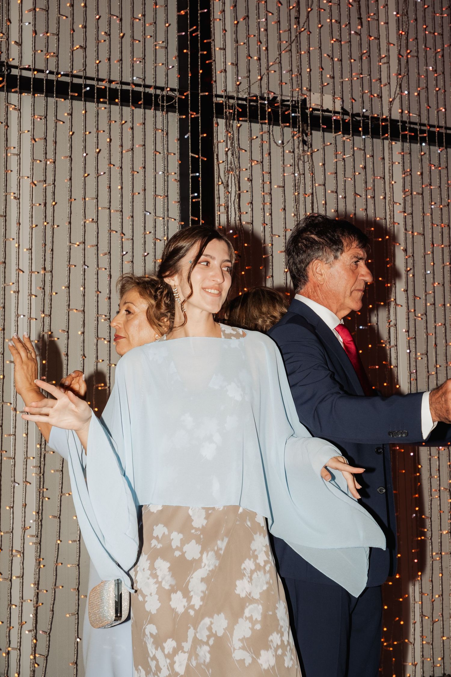 Three elegantly dressed guests posing and dancing in front of a sparkling fairy light backdrop at a wedding.
