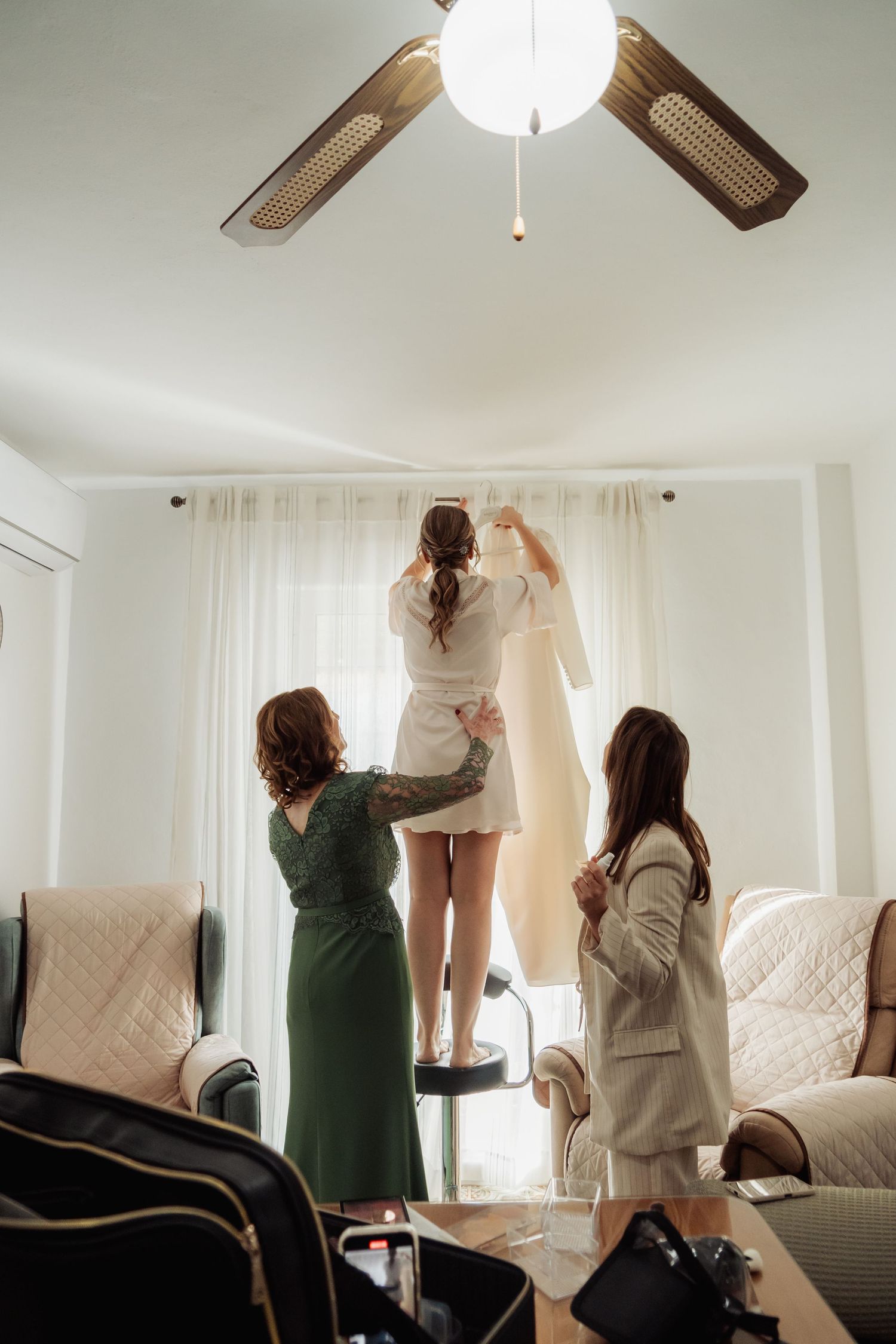 Bride in robe standing on stool reaching for wedding dress while two bridesmaids assist below.