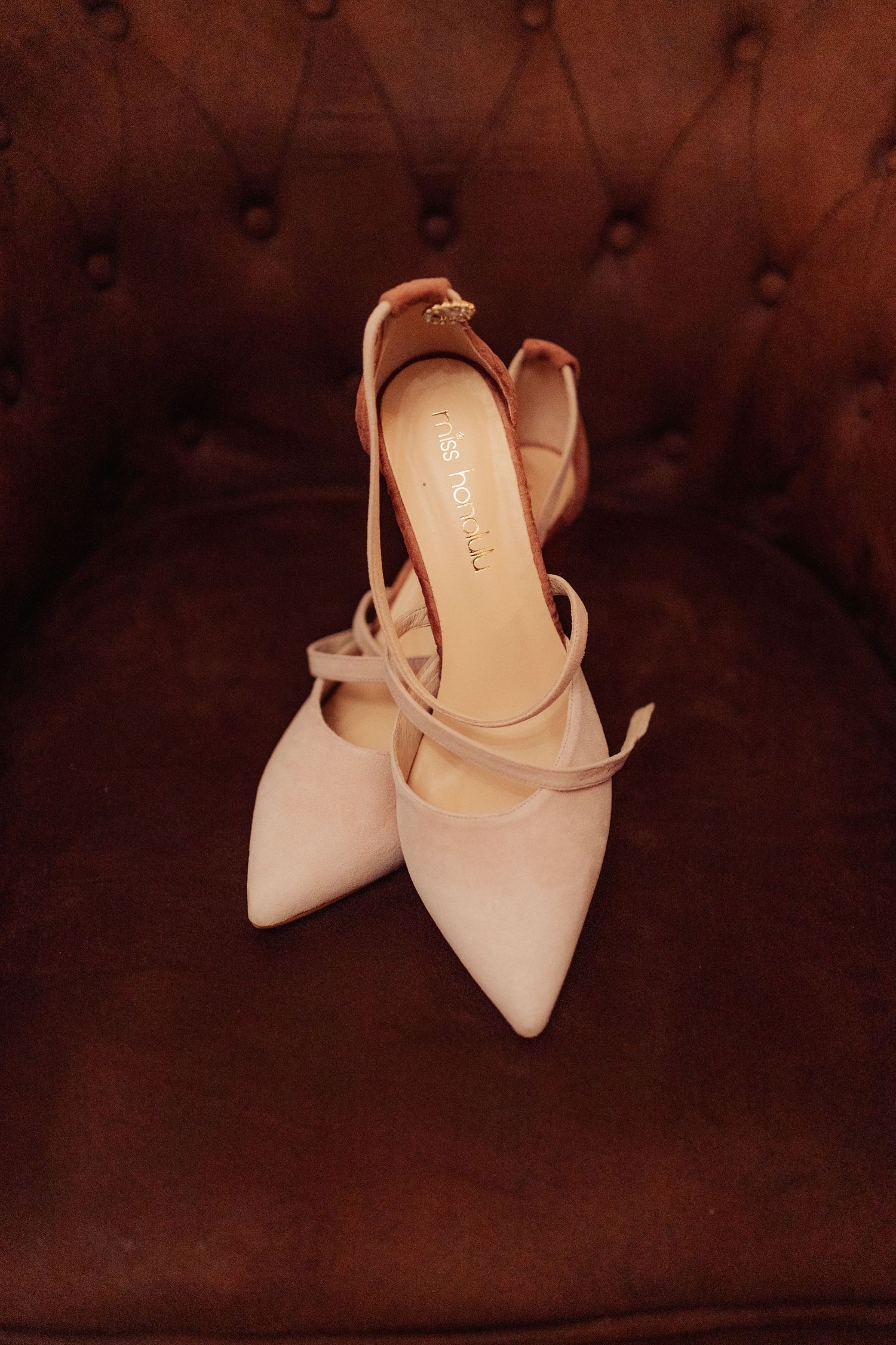 Elegant nude pointed-toe heels with ankle strap detail displayed on brown tufted leather chair.