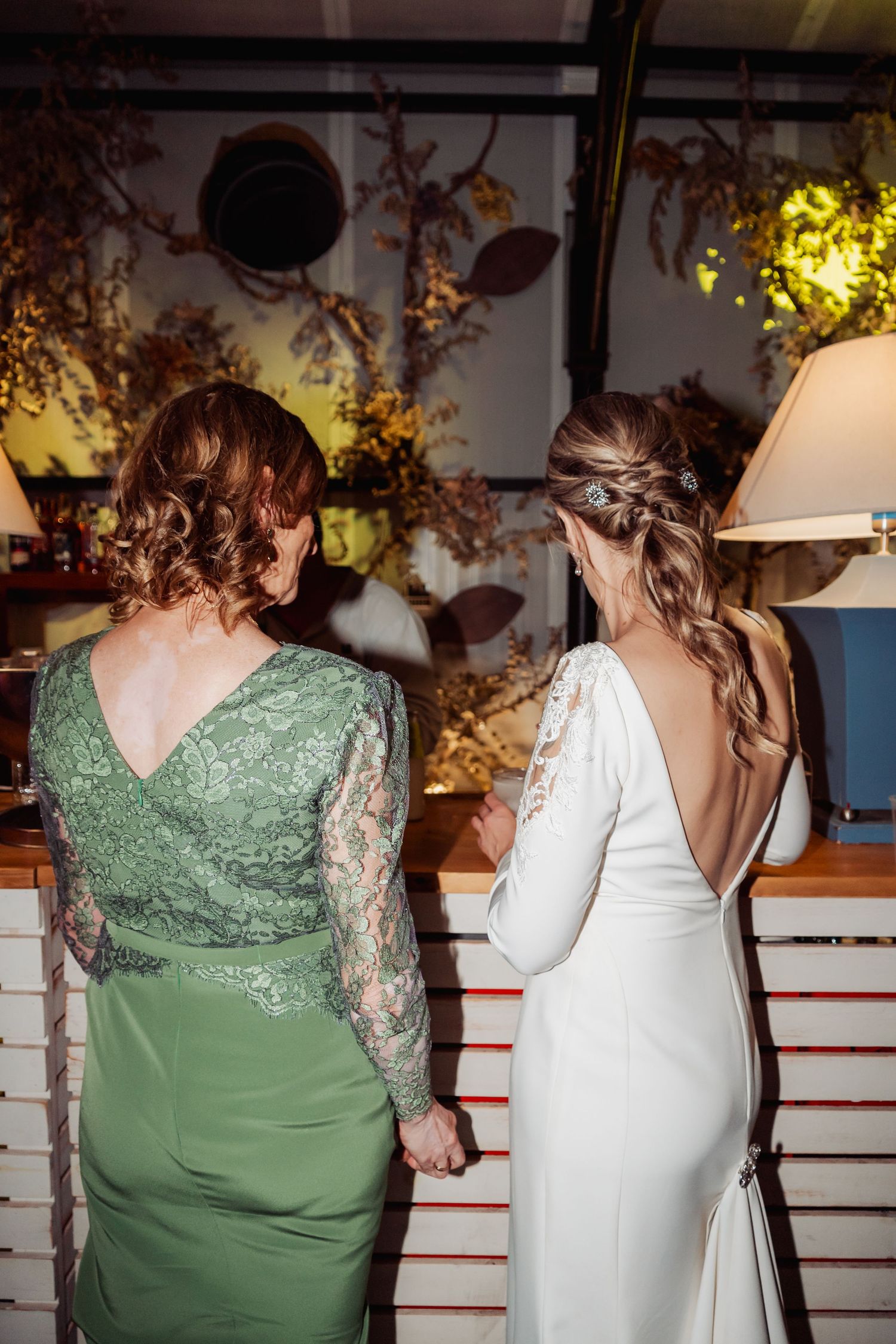 Bride in backless white gown and guest in green lace dress viewed from behind at wedding reception.