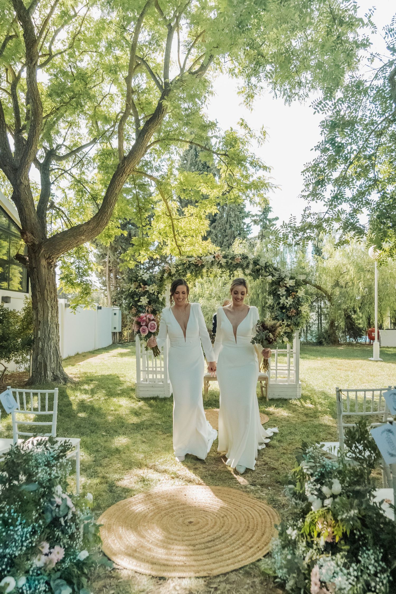 Two brides in white gowns holding hands walking down the aisle at an outdoor garden wedding.