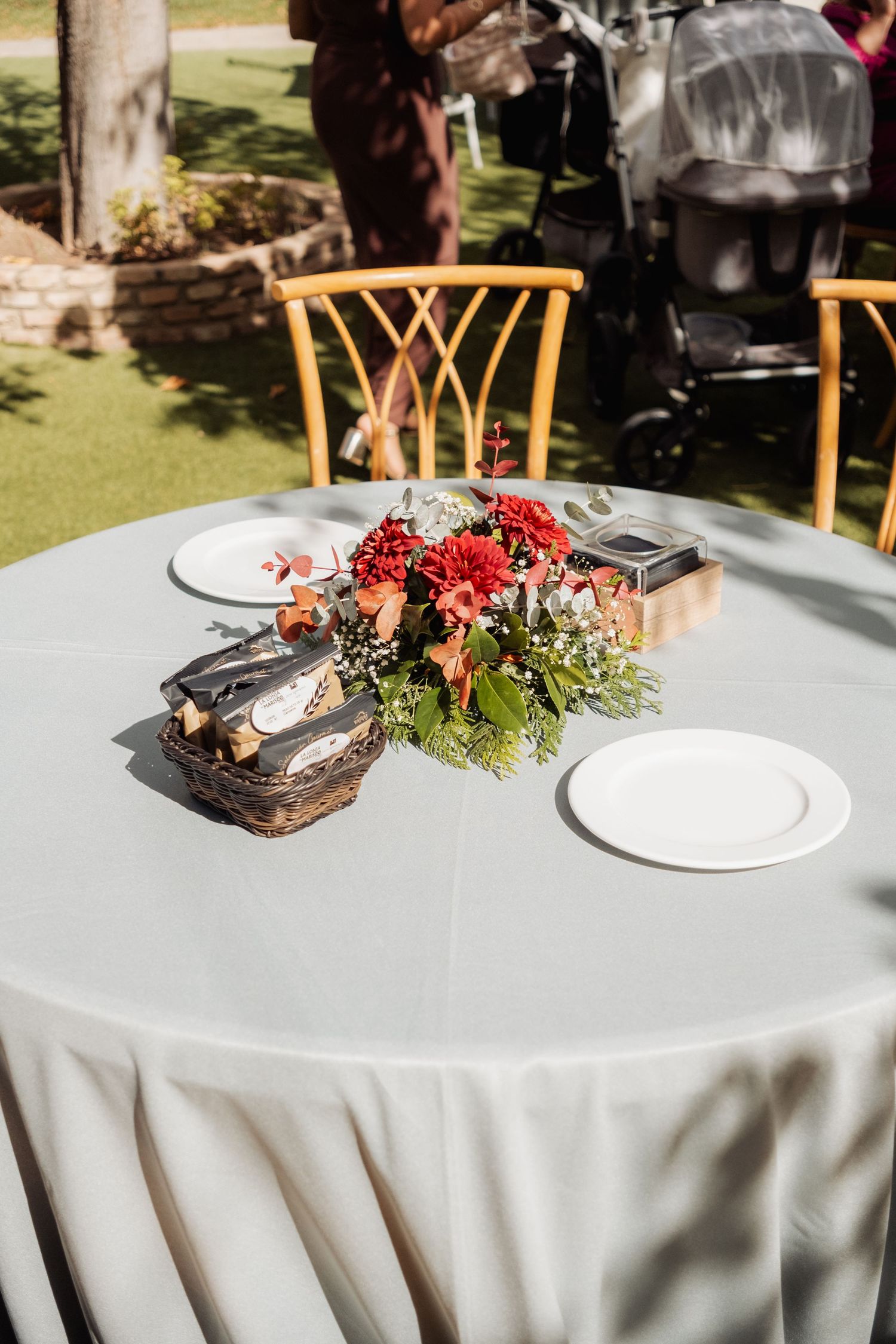 Round outdoor wedding reception table with white linen, white plates, and a red floral centerpiece.